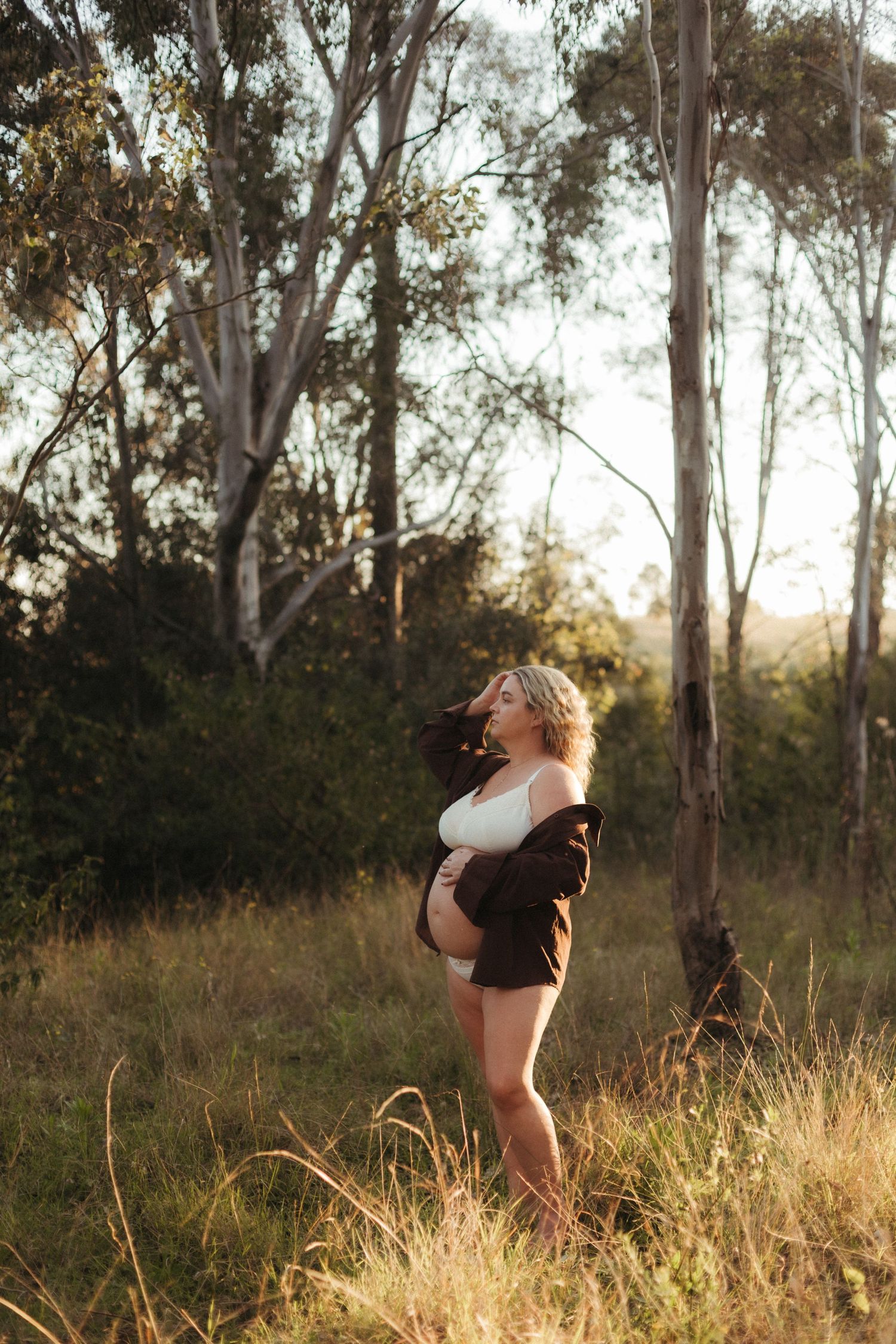 Pregnant woman in white bra and brown jacket standing in golden sunlit meadow.