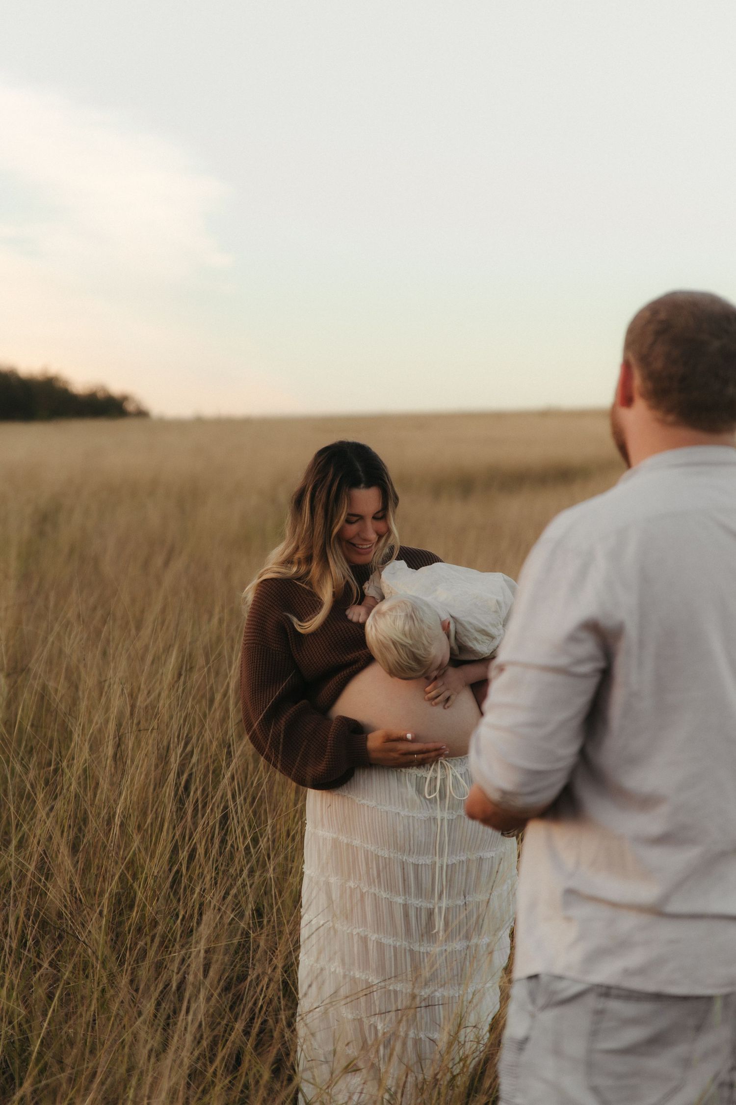 Pregnant woman smiling at baby in golden field at sunset, with man standing nearby, maternity photo shoot.