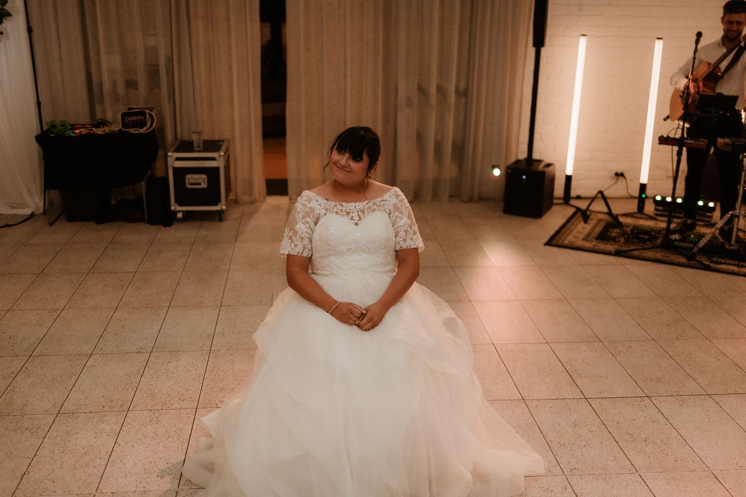 Bride in white lace and tulle wedding gown sits alone on dance floor during wedding reception.