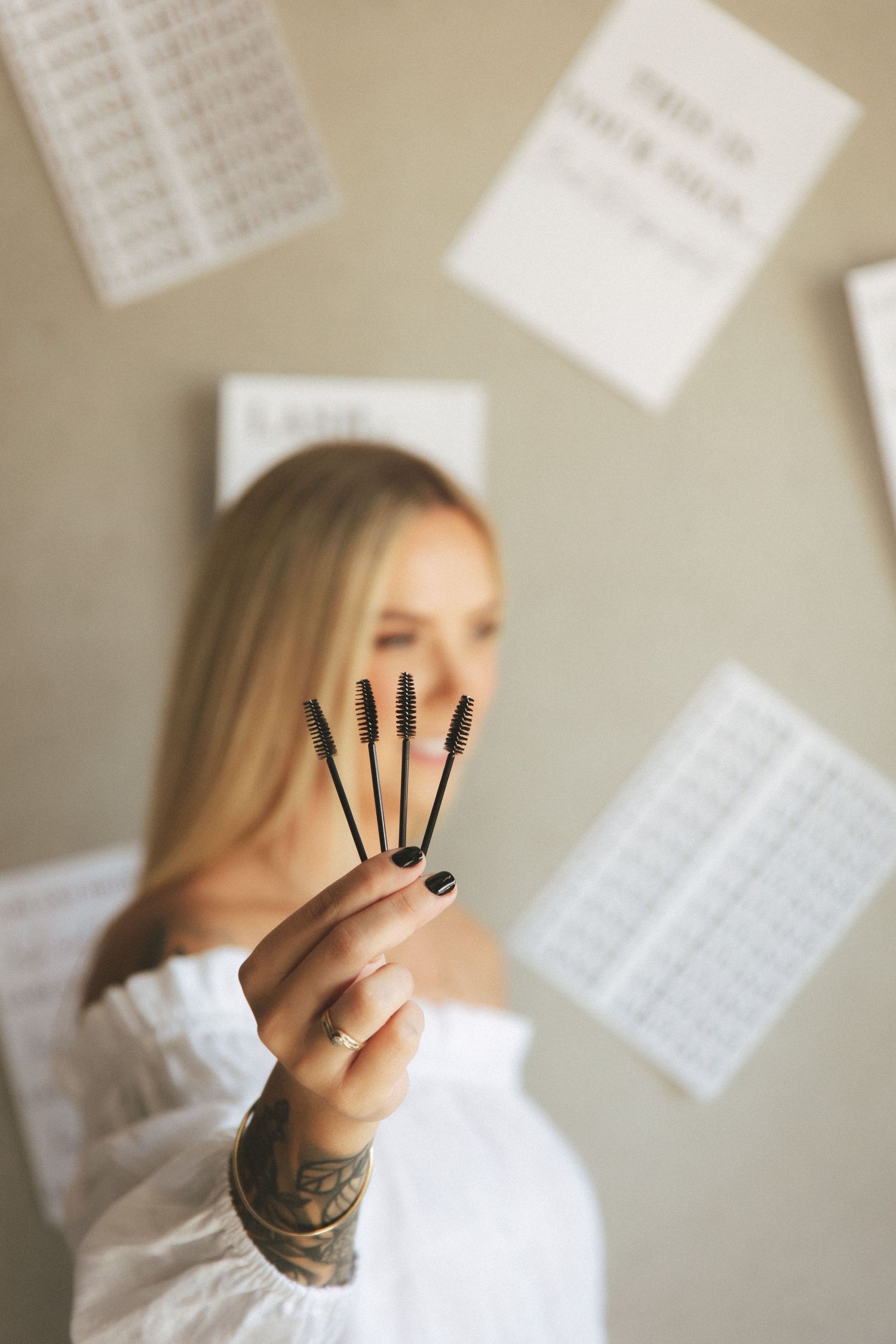 Blonde woman holding four mascara spoolie brushes toward camera in a beauty salon studio.