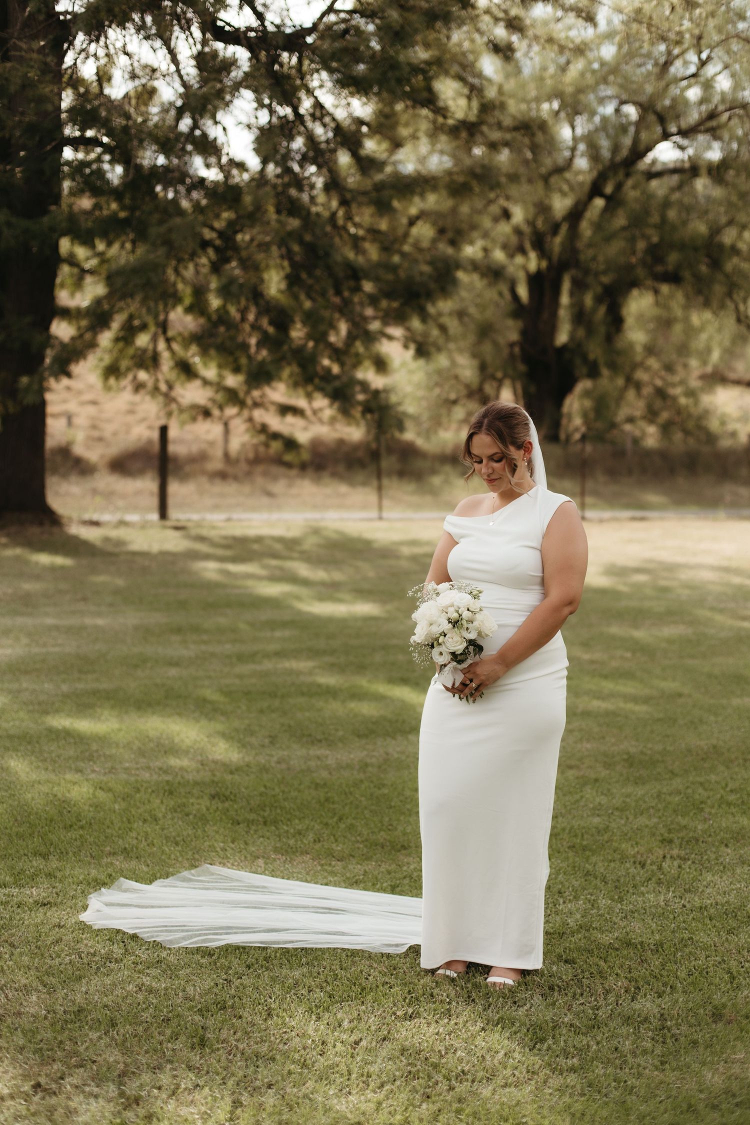 Elegant bride in a fitted white gown with cathedral veil holding a white bouquet in a lush garden.
