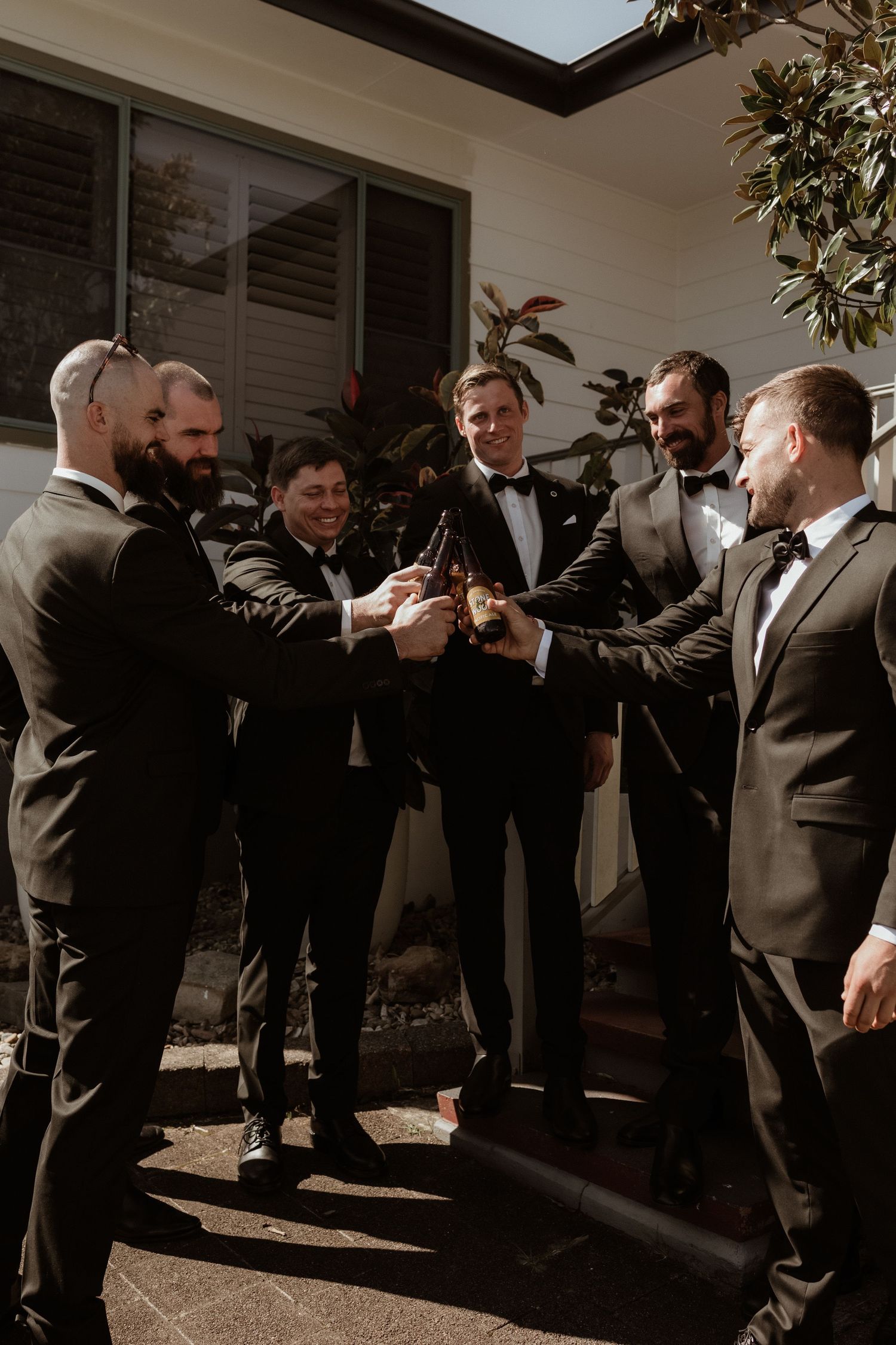 Six groomsmen in black tuxedos toasting with beer bottles outdoors before a wedding ceremony.