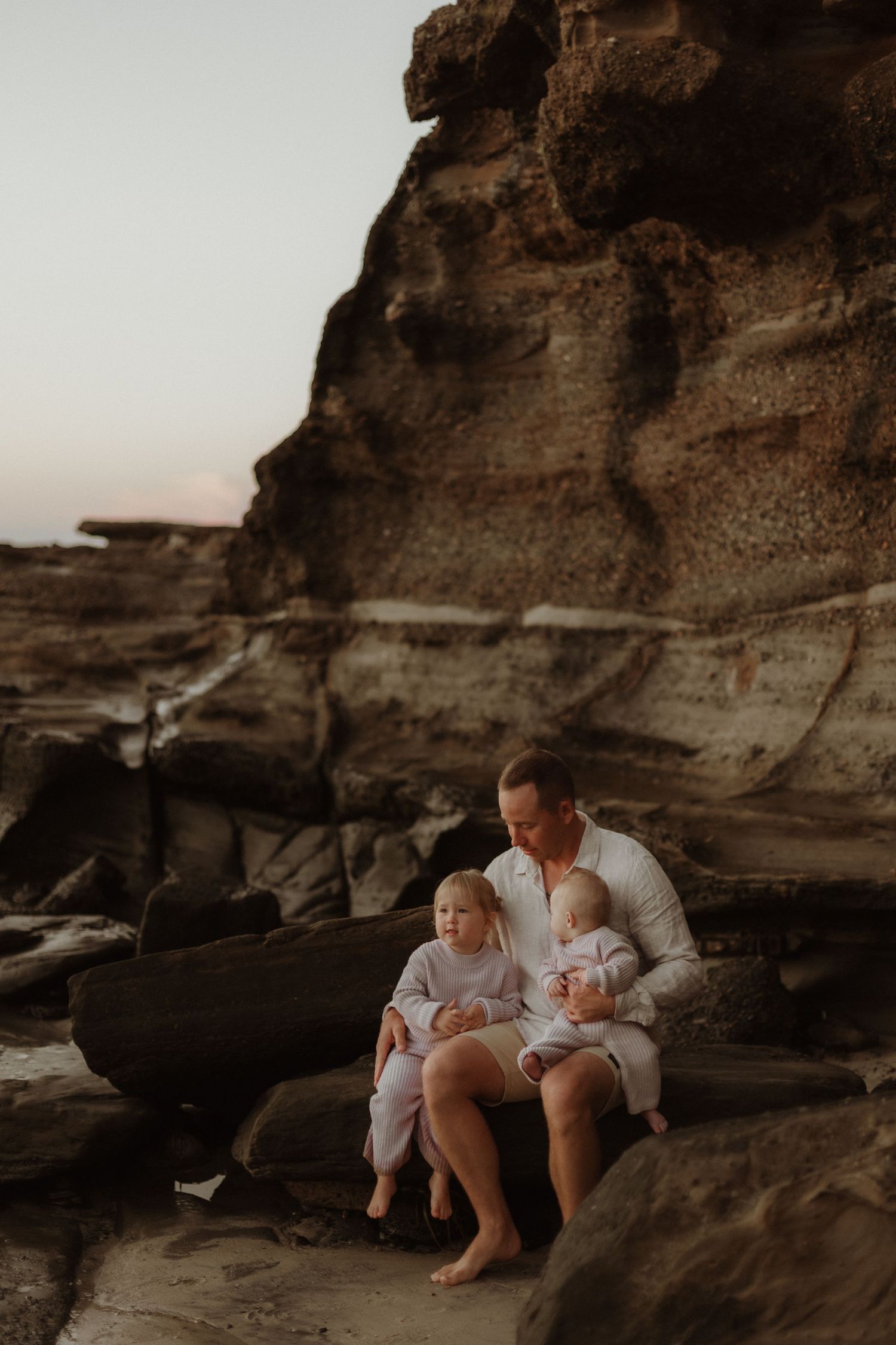 Father sitting on beach rocks cradling two young toddlers near sandstone cliffs at dusk.