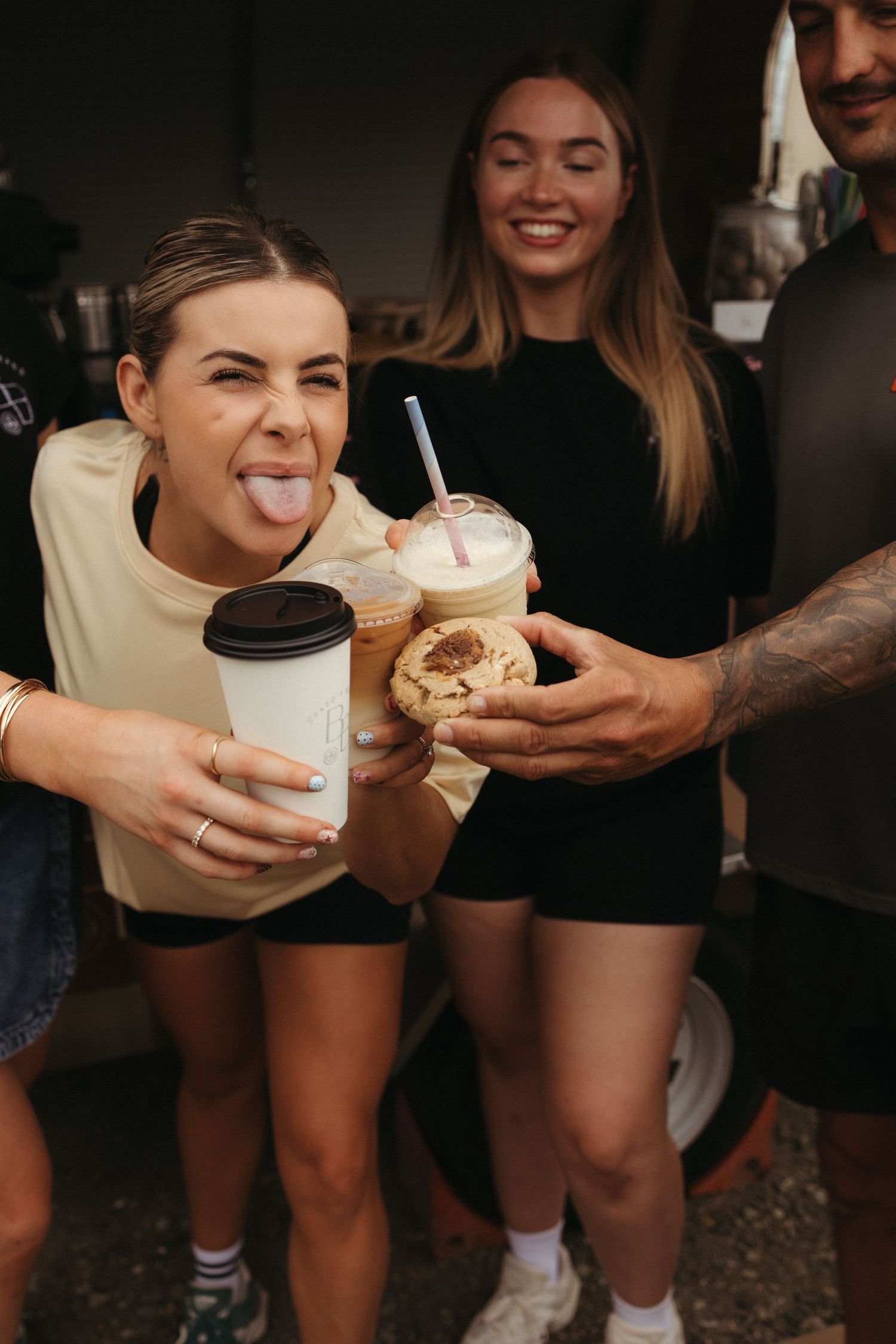 Excited woman sticking out tongue holding coffee cups and cookies with friends outside a coffee van.