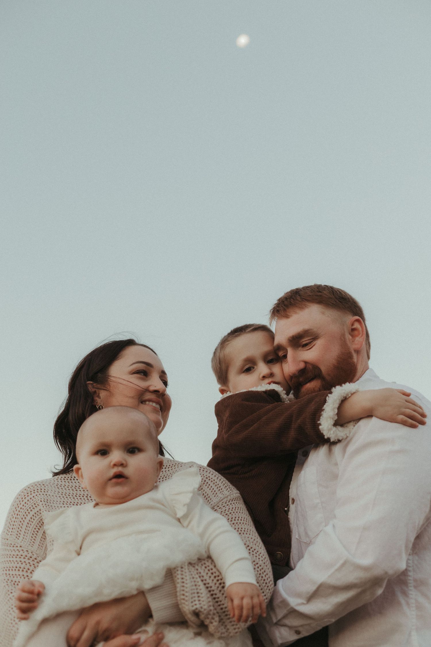 Happy family portrait with parents holding baby and toddler outdoors under a clear evening sky.