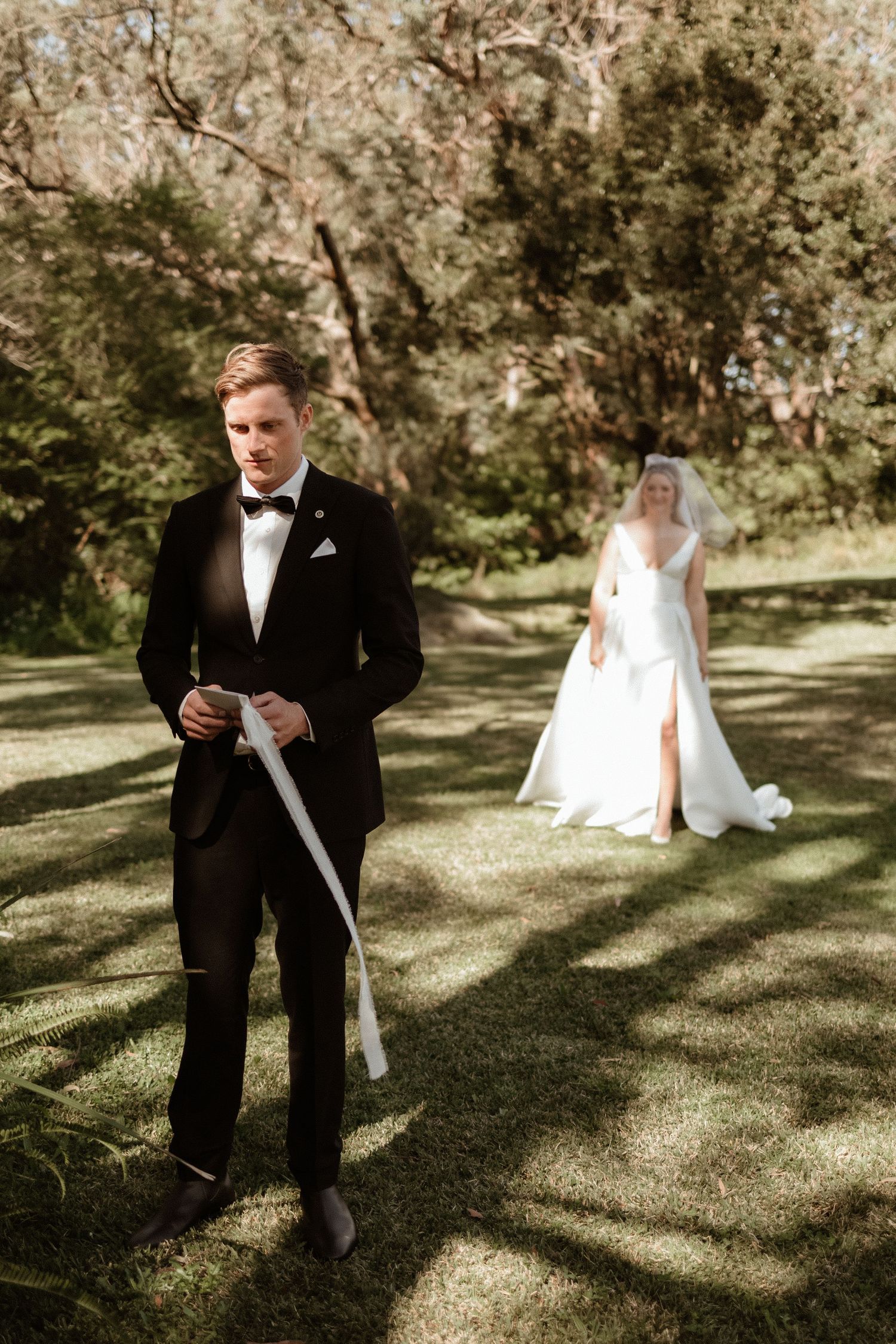 Groom in black tuxedo holds ribbon as bride in white gown approaches through sunlit garden.