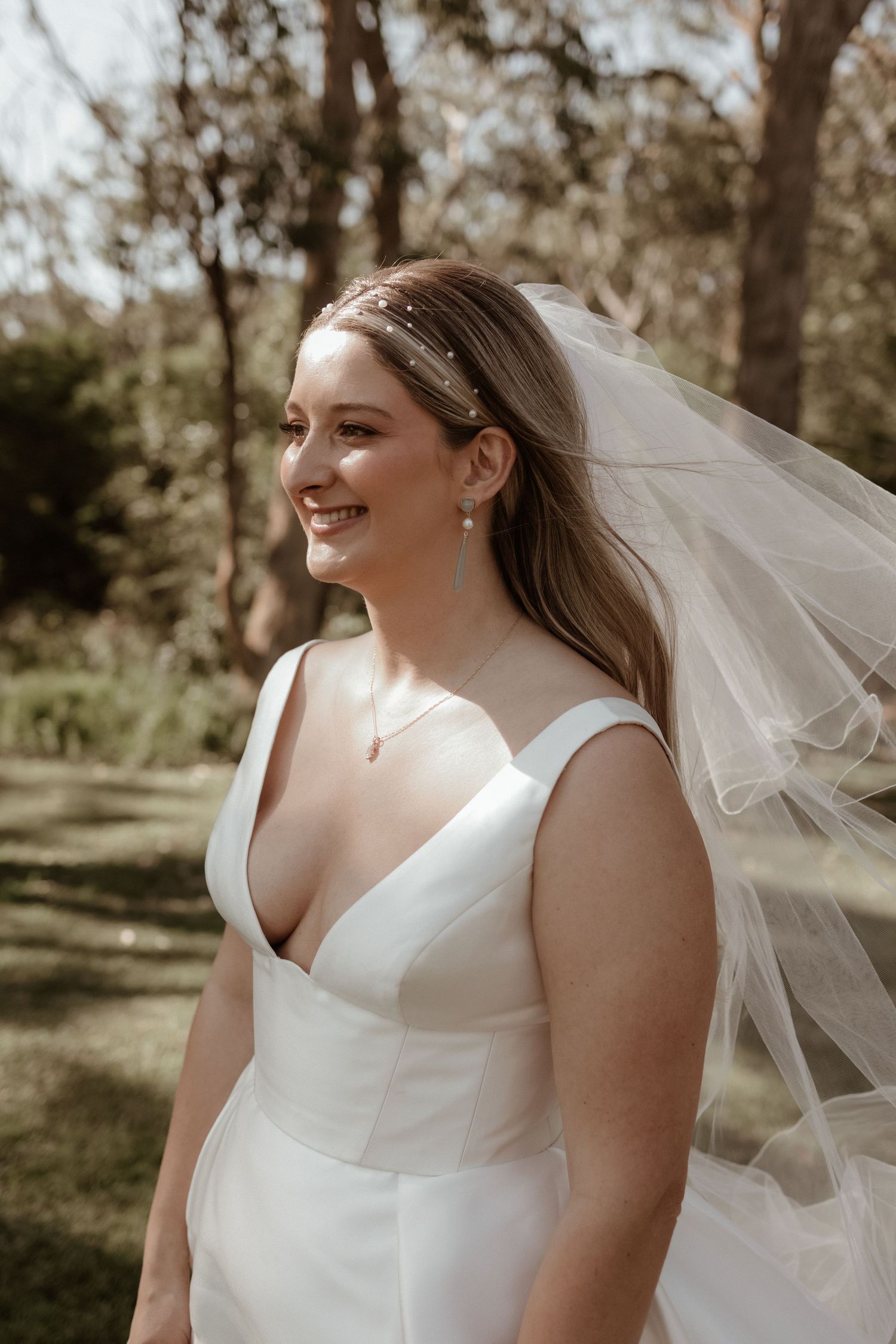 Smiling bride in a white V-neck gown with flowing veil and pearl accessories in a garden setting.