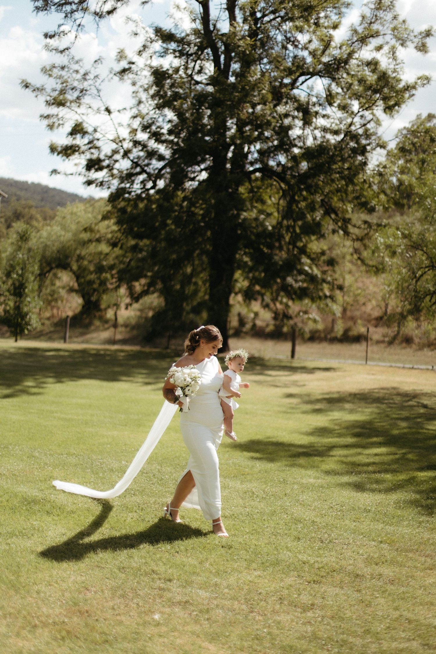 Bride in white dress carrying baby and bouquet walks across lush green lawn with trees in background.