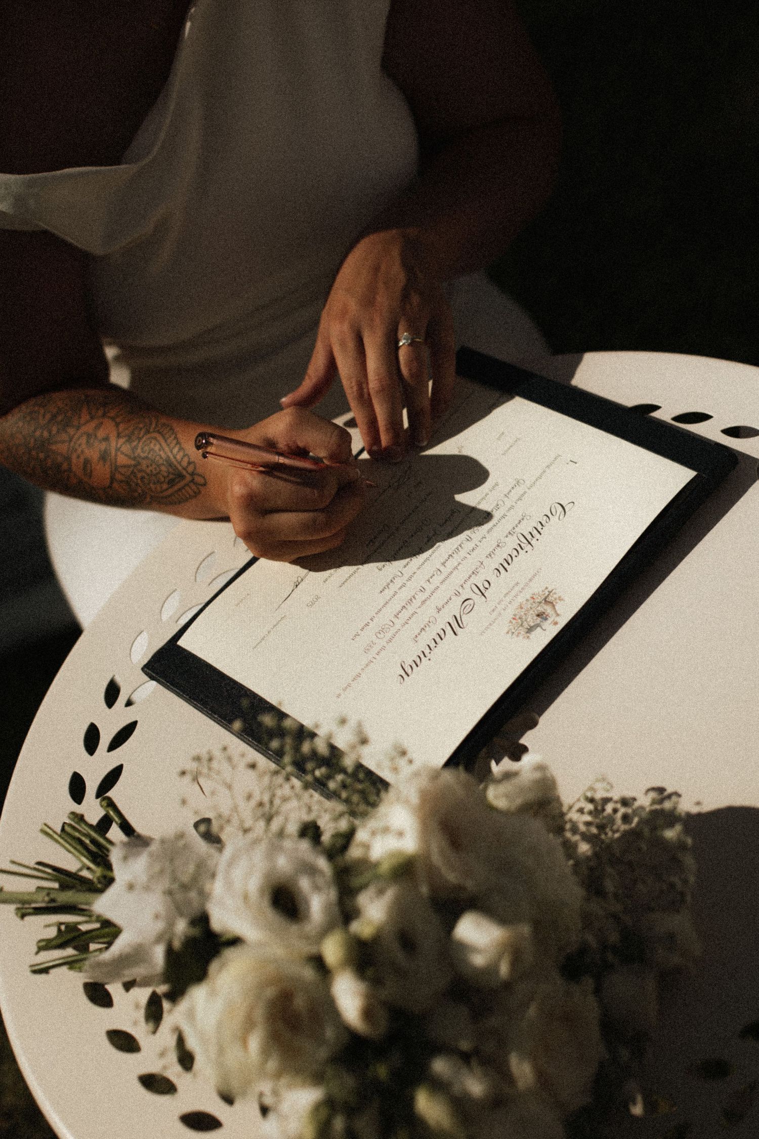 Couple signing a framed marriage certificate on a white table with white floral bouquet nearby.
