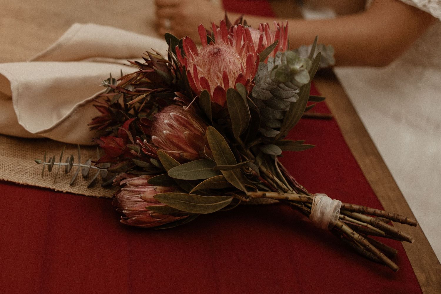 Bridal bouquet featuring pink king protea and native Australian foliage on a burgundy table runner.