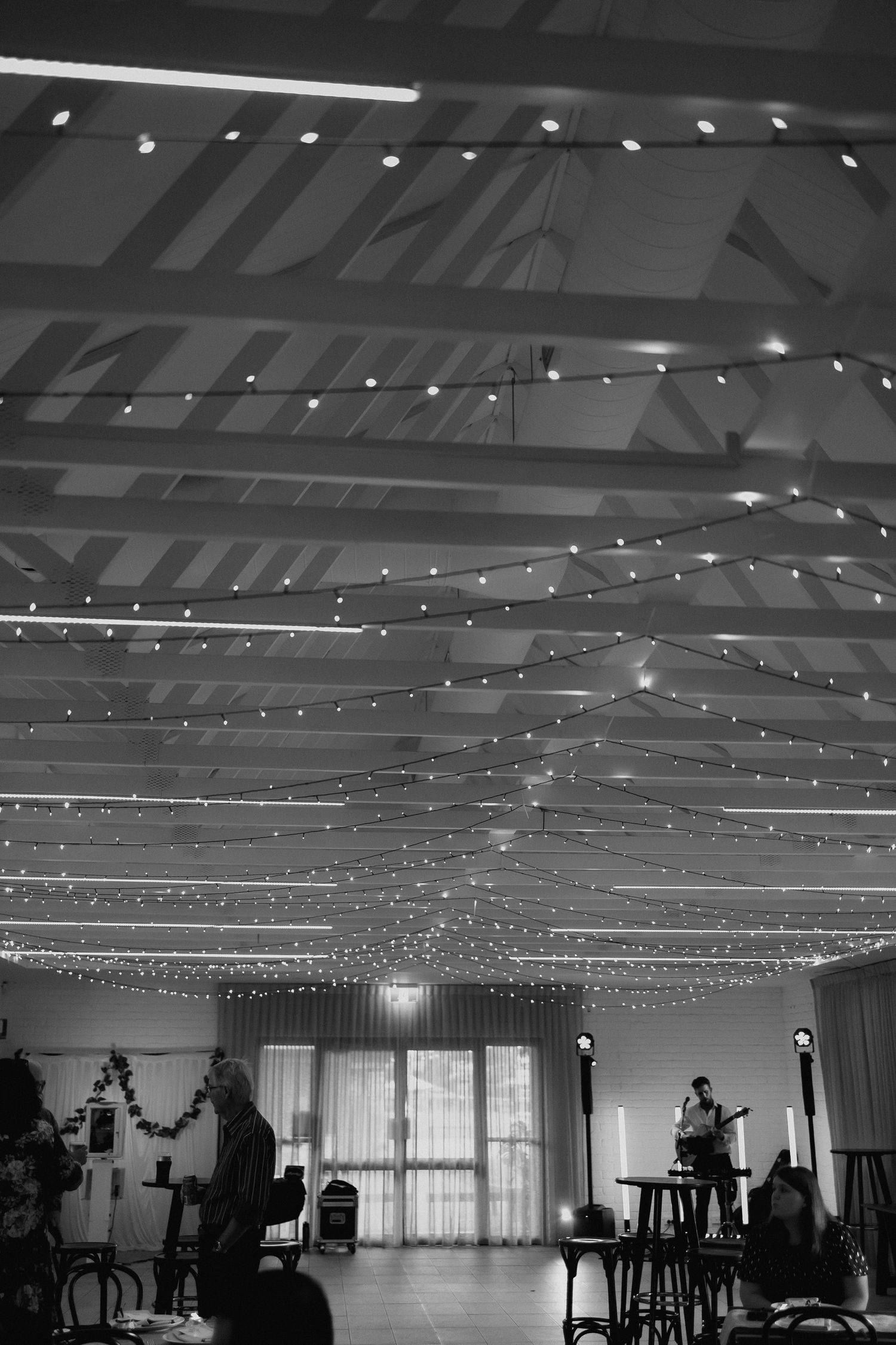 Black and white photo of wedding reception hall ceiling draped with glowing fairy light strands.