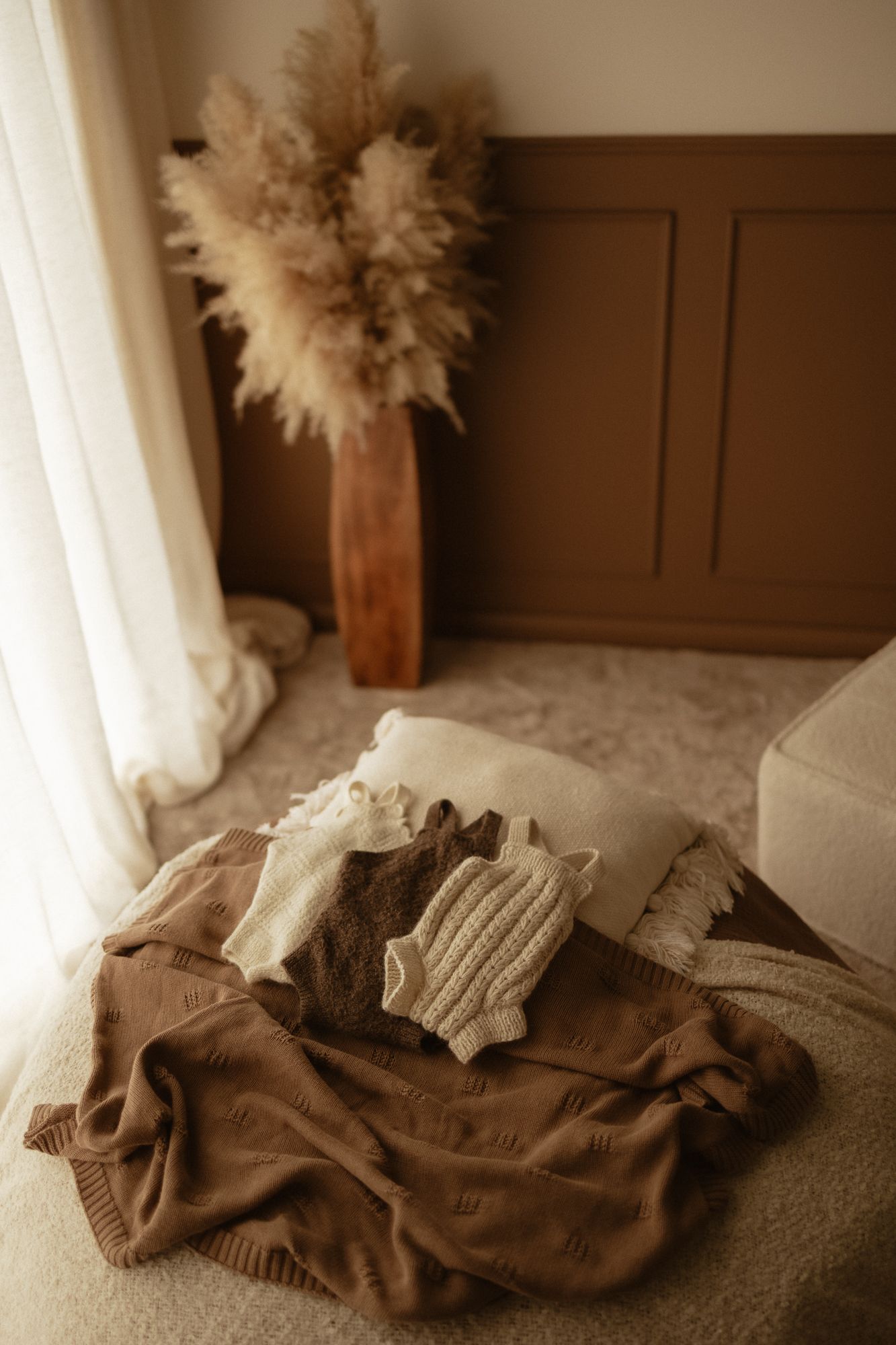 Cozy brown knit blankets and cream sweaters stacked on a fluffy rug beside pampas grass in a warm boho room.