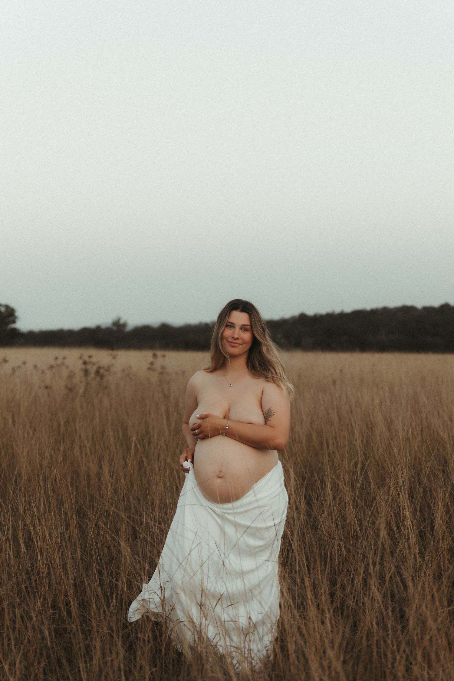 Pregnant woman in a field of tall golden grass, draped in a white cloth, cradling her baby bump at dusk.