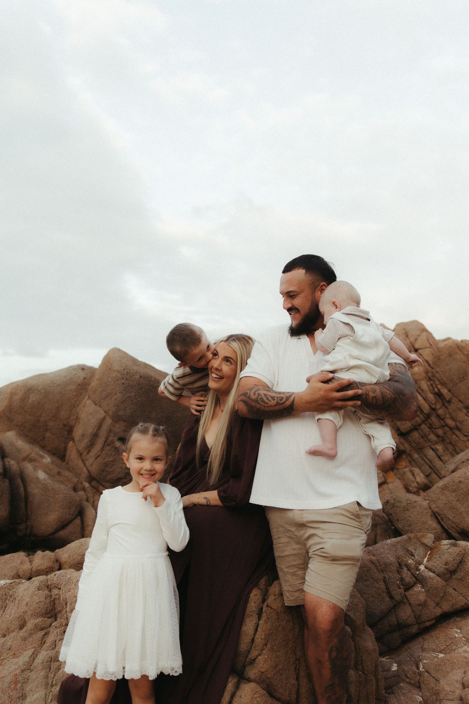 A happy family of five poses on rocky beach, parents holding two babies while young girl in white dress smiles.