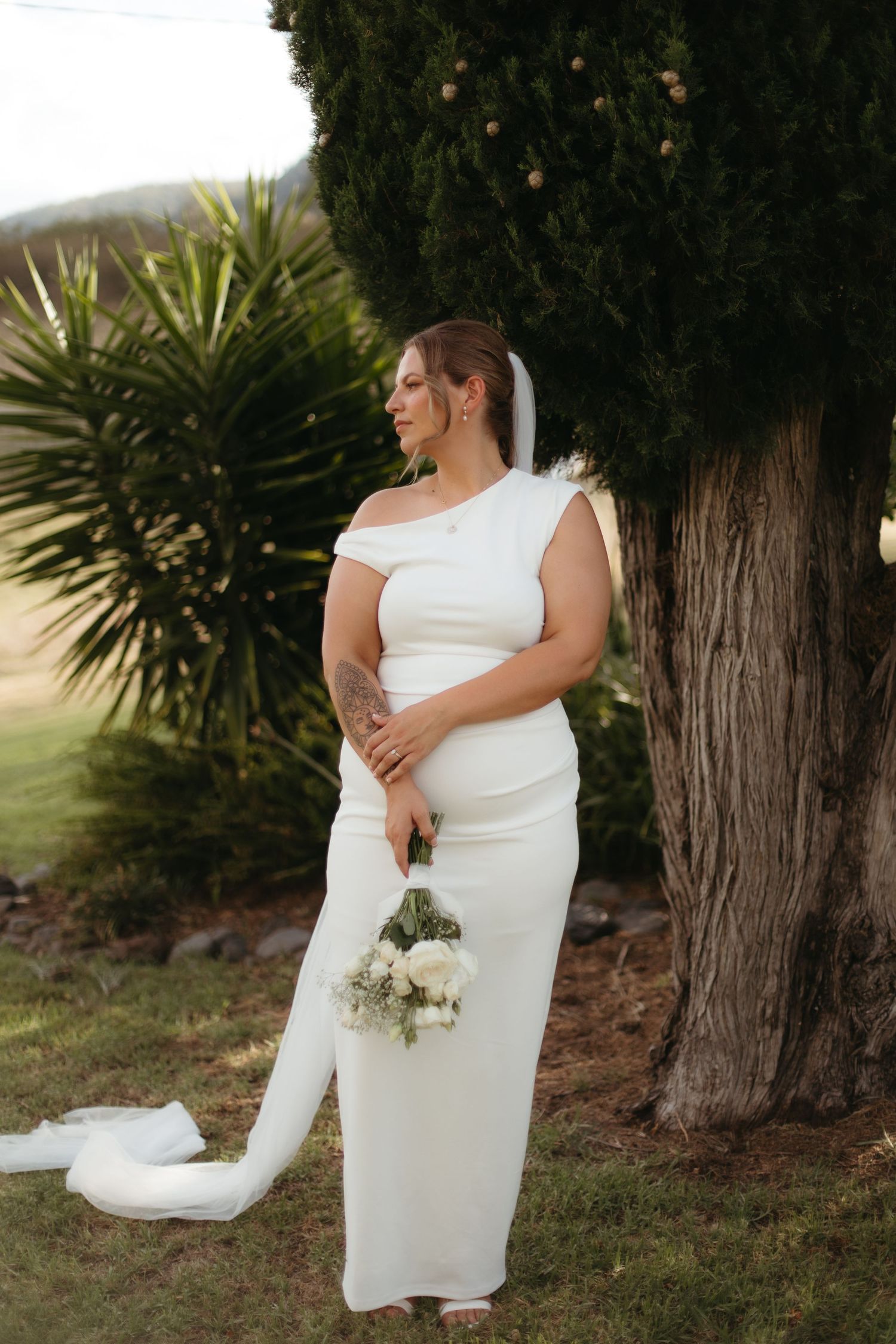 Beautiful bride in white fitted gown standing beside tree holding white floral bouquet.