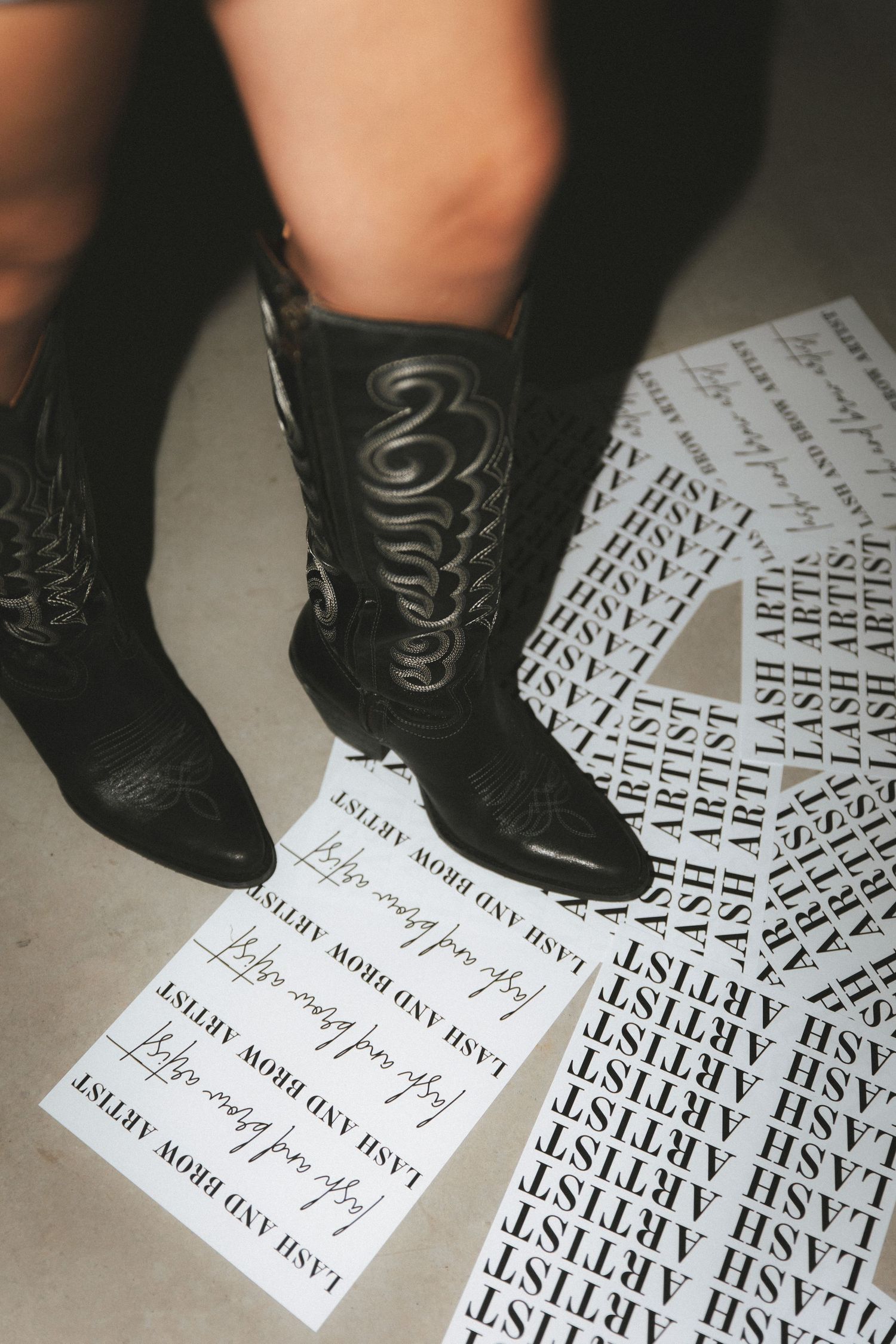 Close-up of black cowboy boots surrounded by scattered Lash Artist and lash brow artist printed papers.