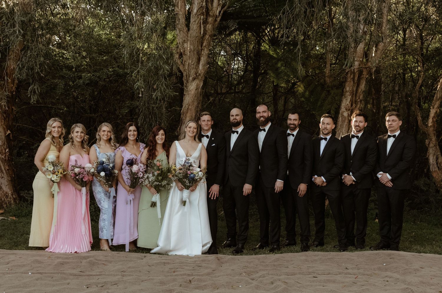 Wedding party posing in colorful mismatched bridesmaid dresses and black tuxedos in a lush forest setting.