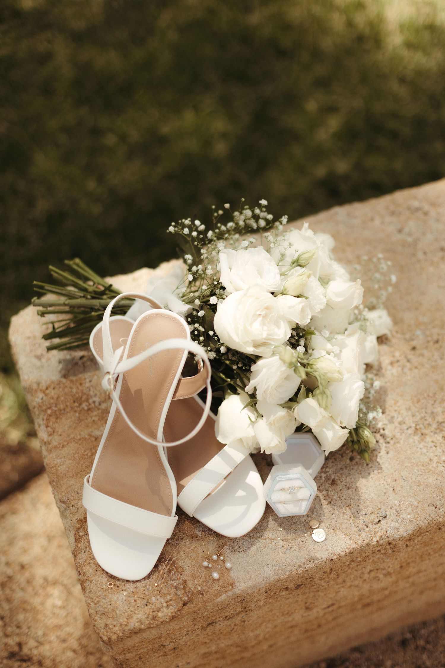 White bridal heels, bouquet of white roses, and ring box displayed on stone surface.