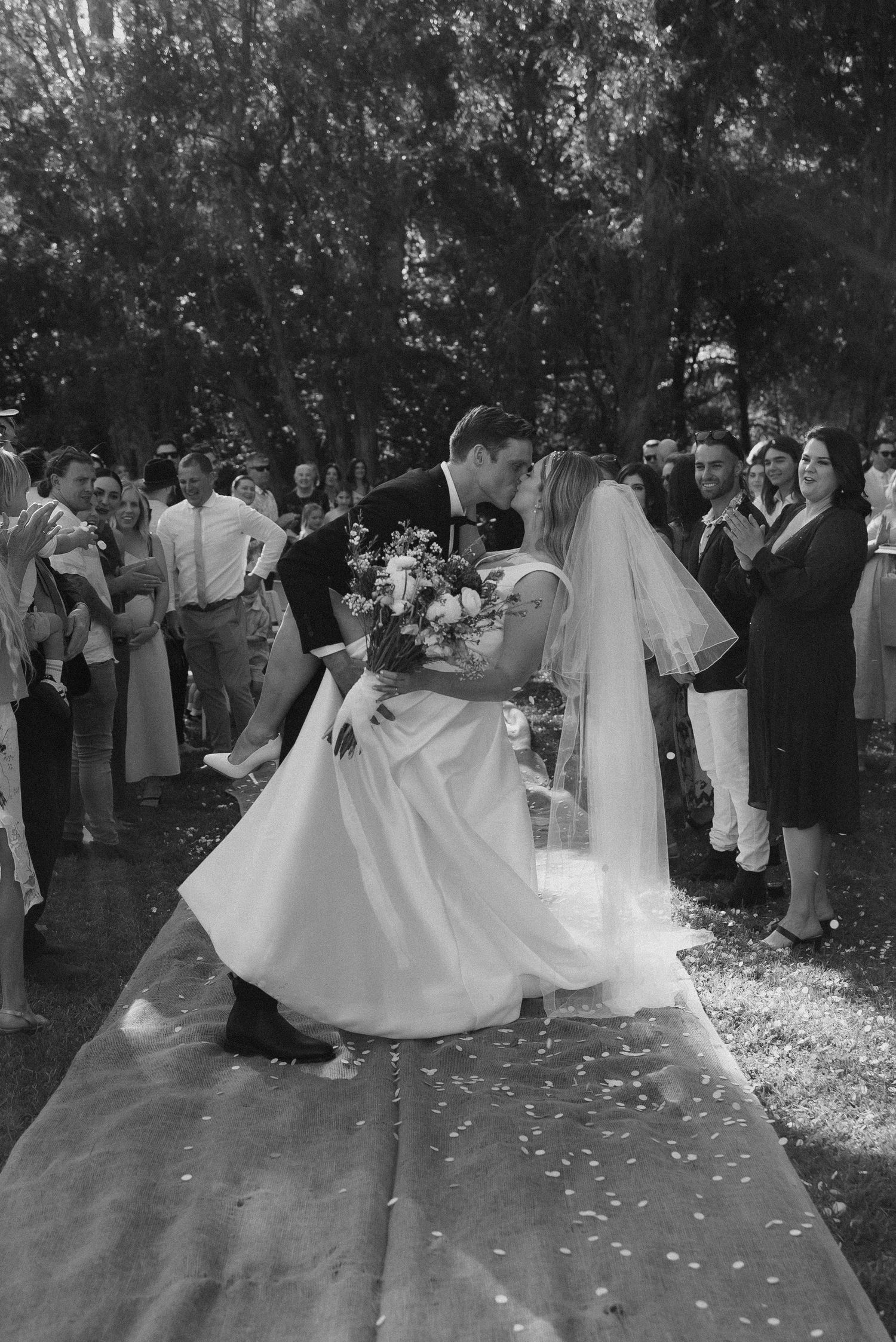 Black and white photo of a bride and groom sharing their first kiss at an outdoor wedding ceremony.