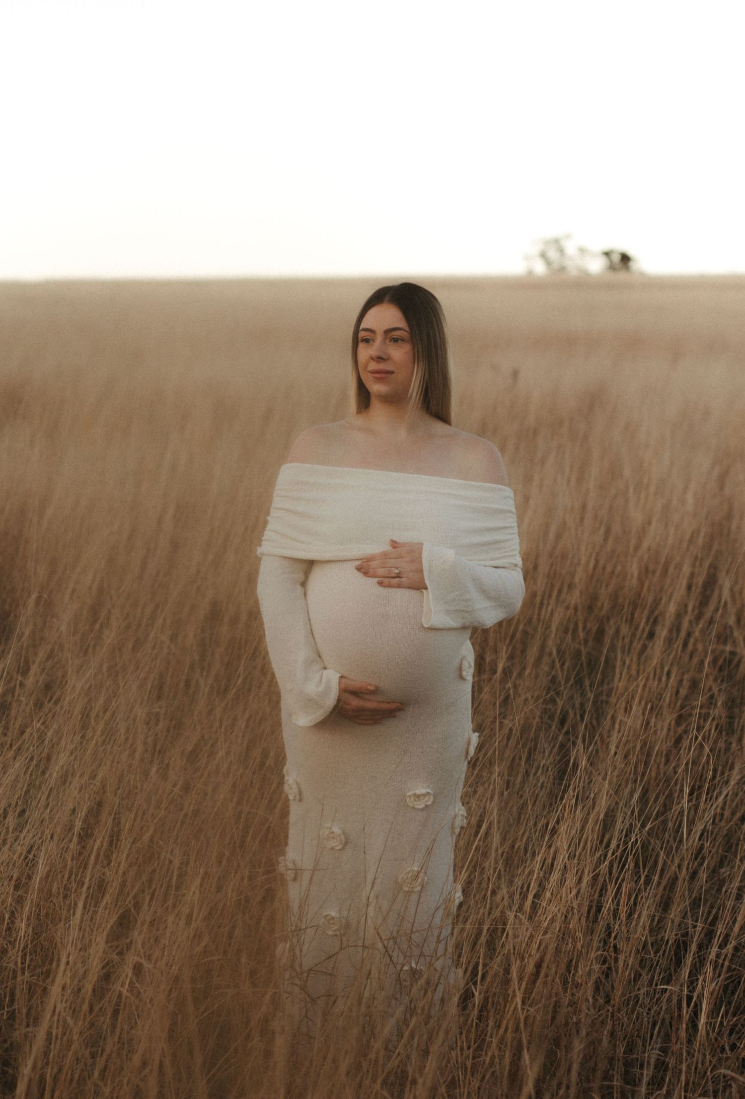 Pregnant woman in off-shoulder white dress cradling her bump while standing in a golden wheat field.