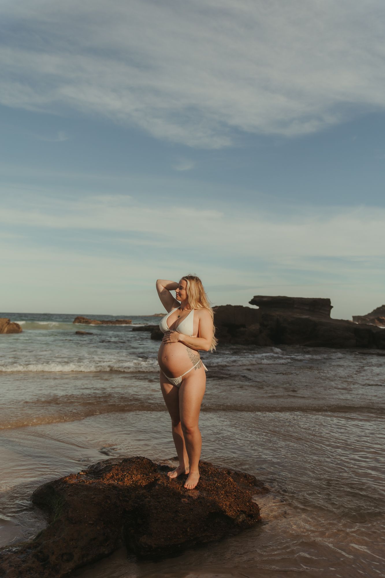 Pregnant woman in white bikini standing on beach rock with ocean waves and rocky shore in background.