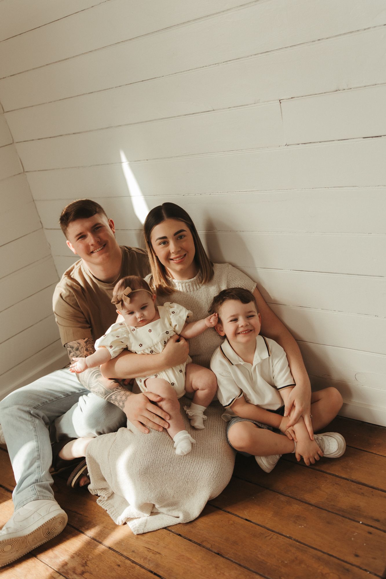 Happy young family with two children sitting on wooden floor against white shiplap wall in cozy home portrait.