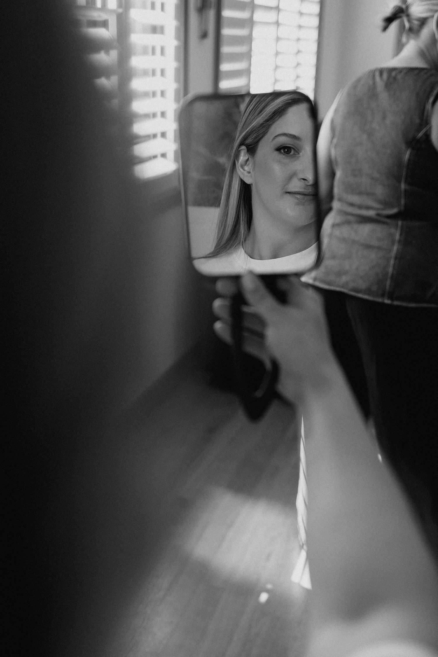 Black and white reflection of a smiling blonde bride in a handheld mirror during wedding preparations.
