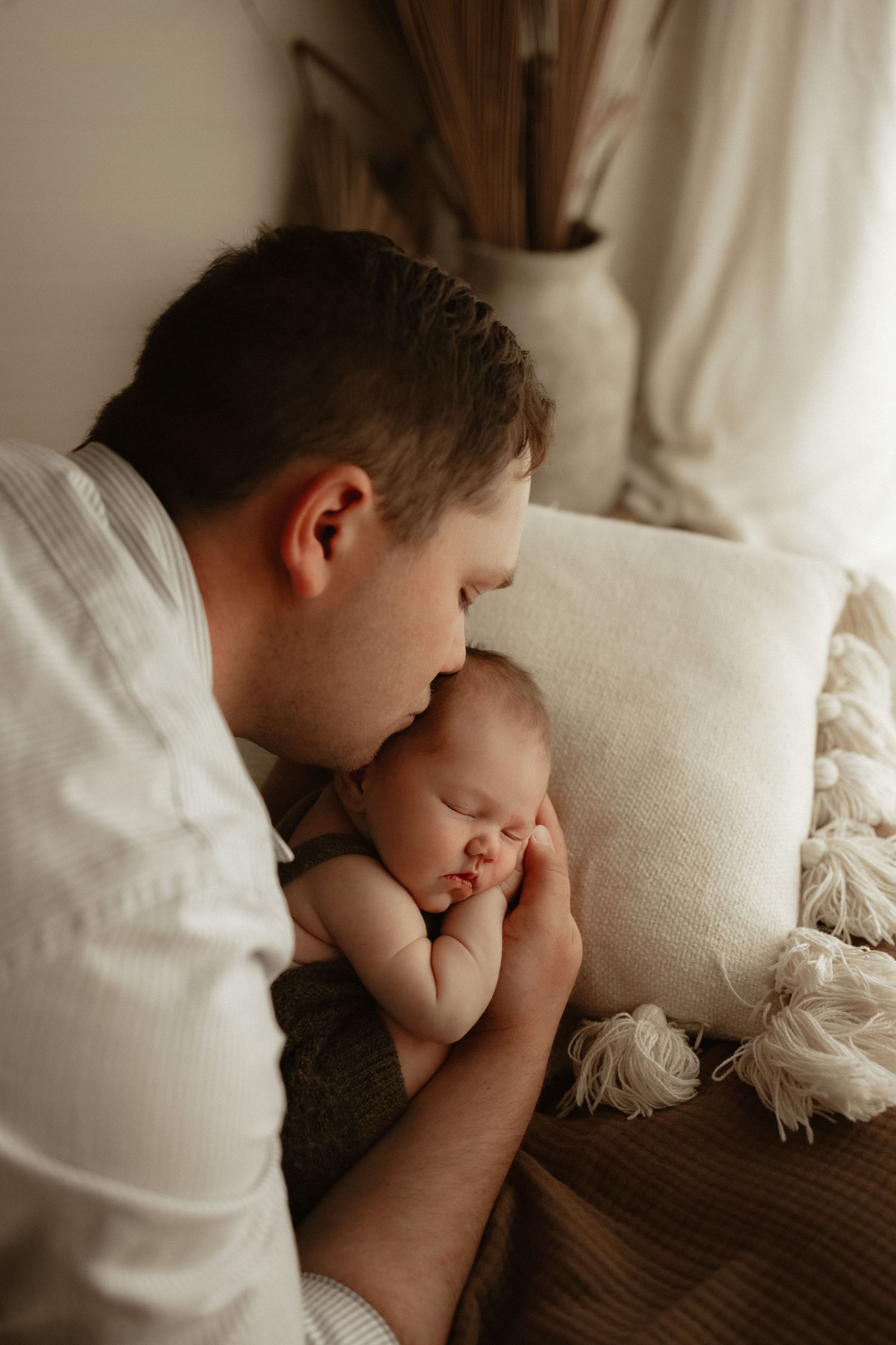 A tender father gently kisses his sleeping newborn baby resting on a soft blanket with neutral boho decor.
