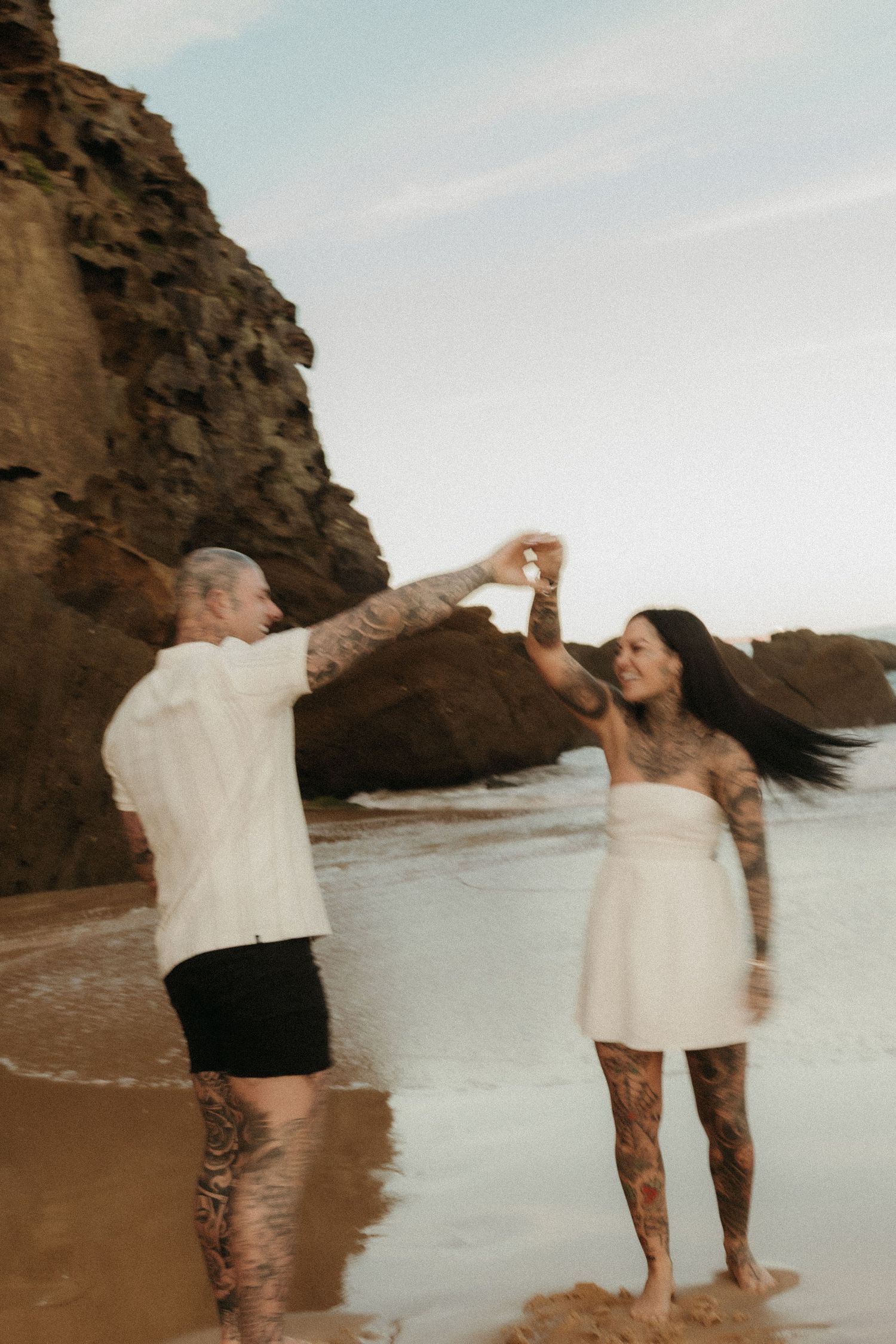 Tattooed couple dancing on beach near rocky cliffs with woman spinning in white dress.