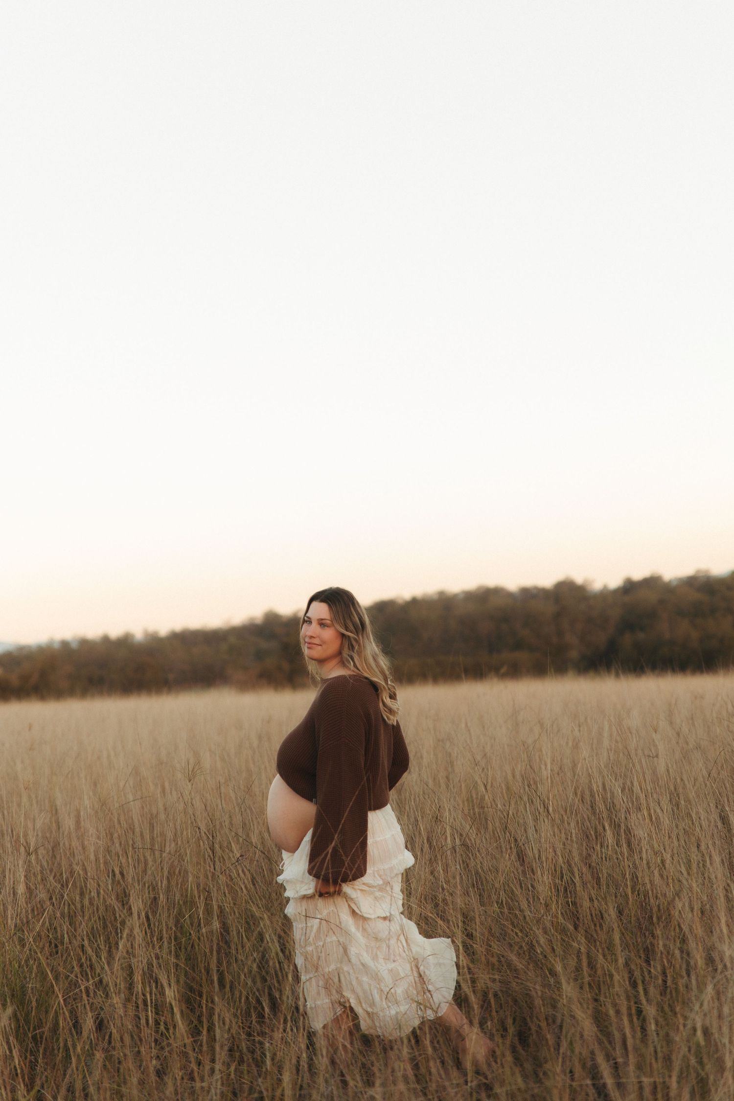 Pregnant woman in brown top and white skirt standing in golden field at sunset, looking over shoulder.