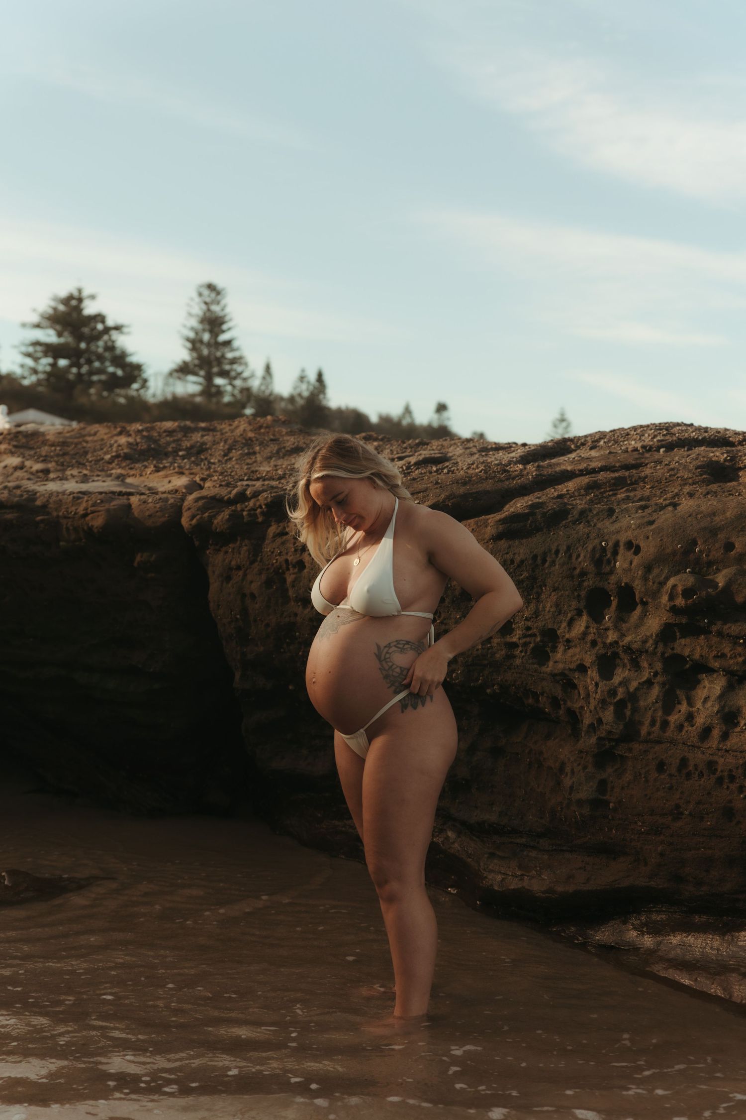 Pregnant woman in white bikini standing among coastal rocks with waves at her feet at sunset.