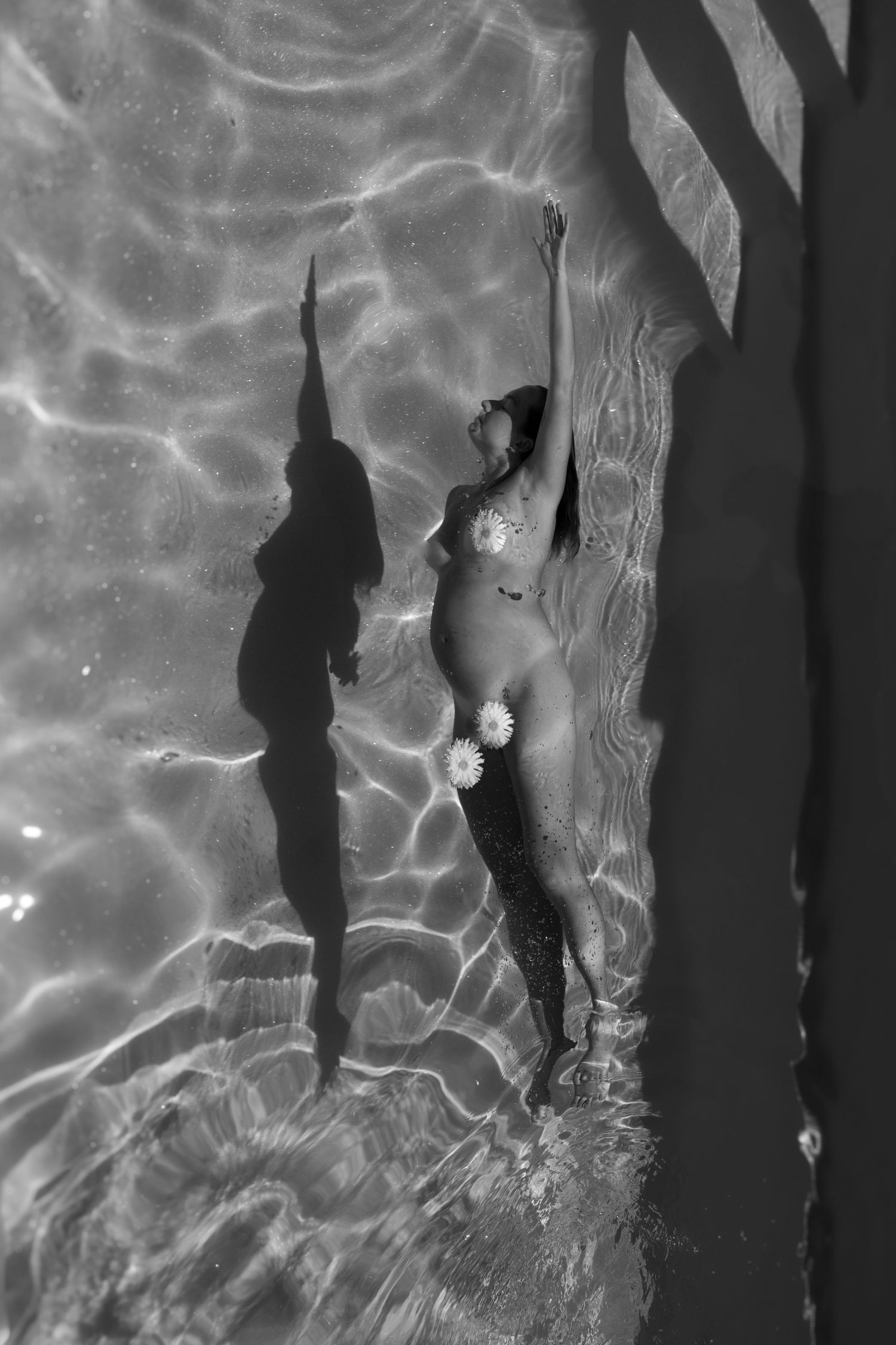 Black and white photo of a nude pregnant woman standing in a pool with daisy flowers.