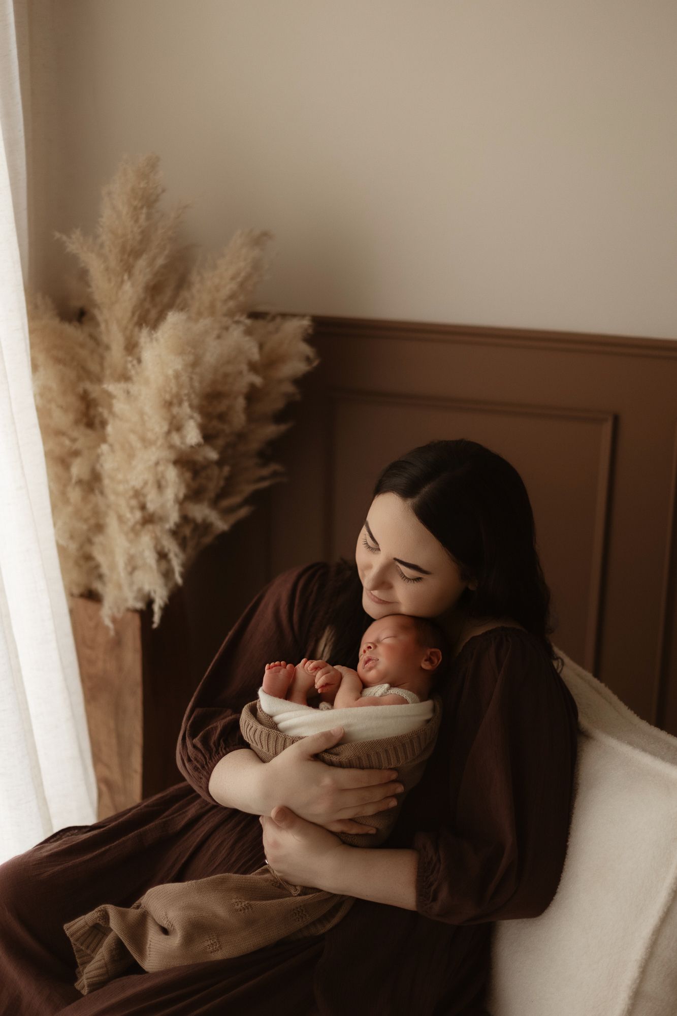 A mother in a brown dress tenderly holds her sleeping newborn baby in a warm, neutral-toned room with pampas grass decor.