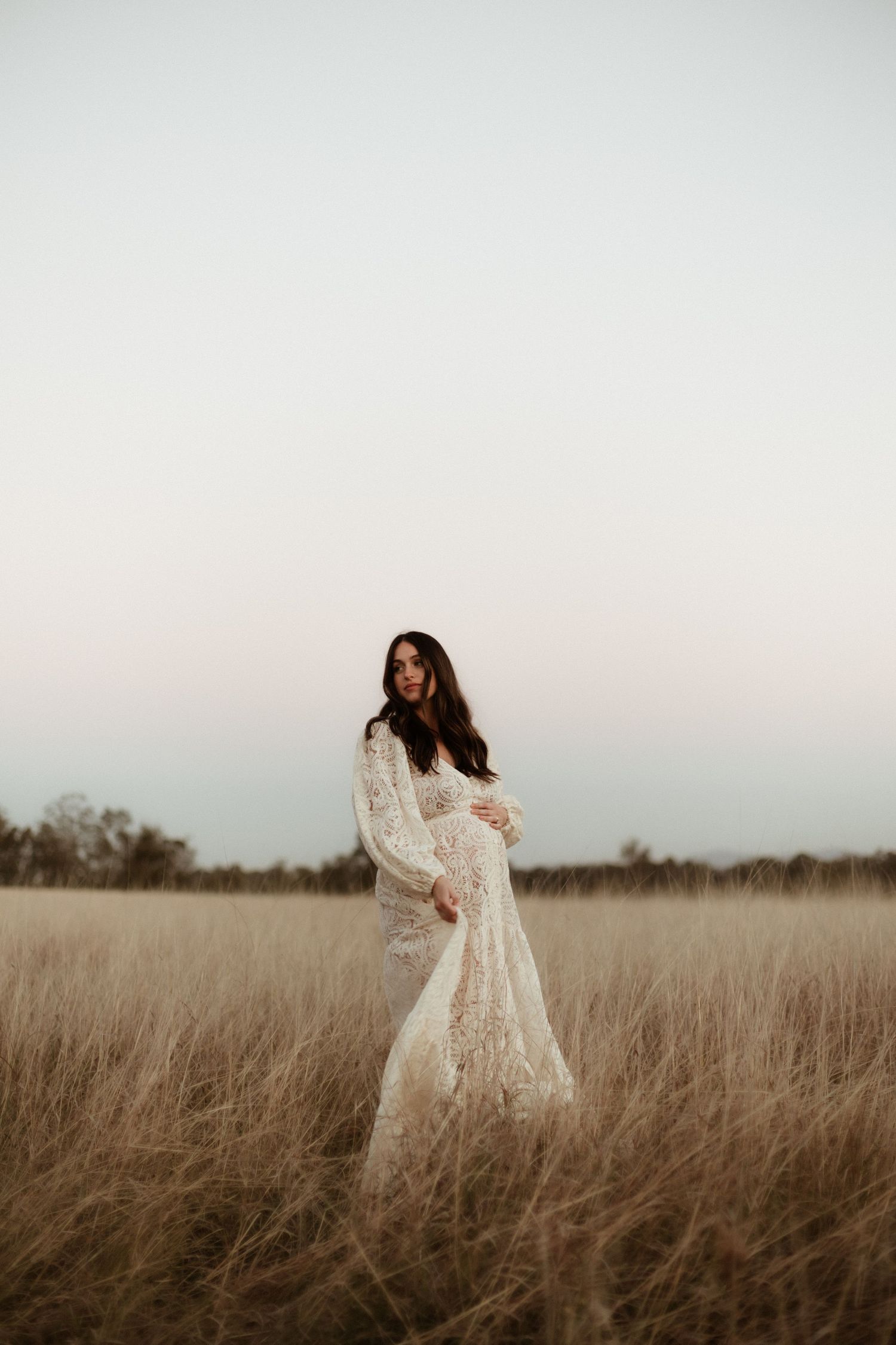 Pregnant woman in flowing white lace dress standing in golden field at twilight.