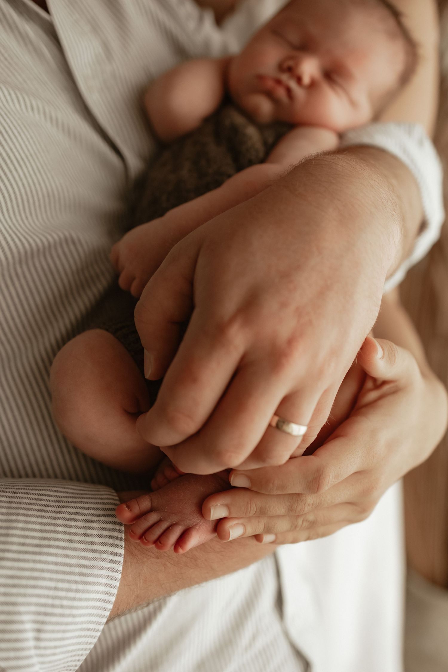 dad cradling a sleeping newborn baby, with tiny feet visible and a wedding ring on one hand. warm soft, neutral authentic emotive brown