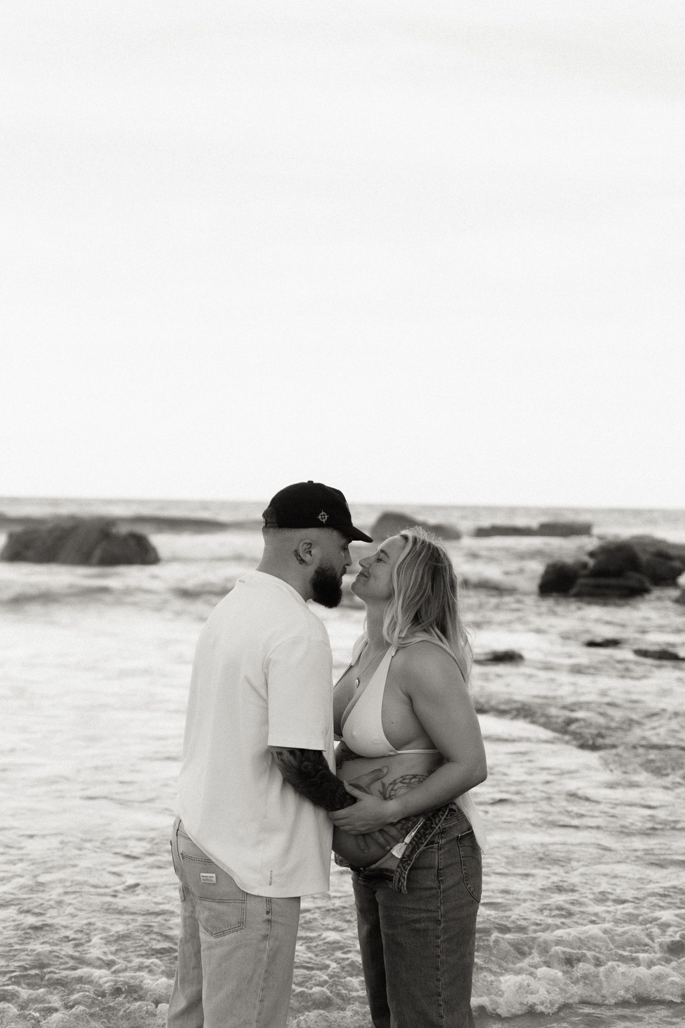 Black and white photo of a couple sharing a romantic kiss on a rocky beach with ocean waves in the background.