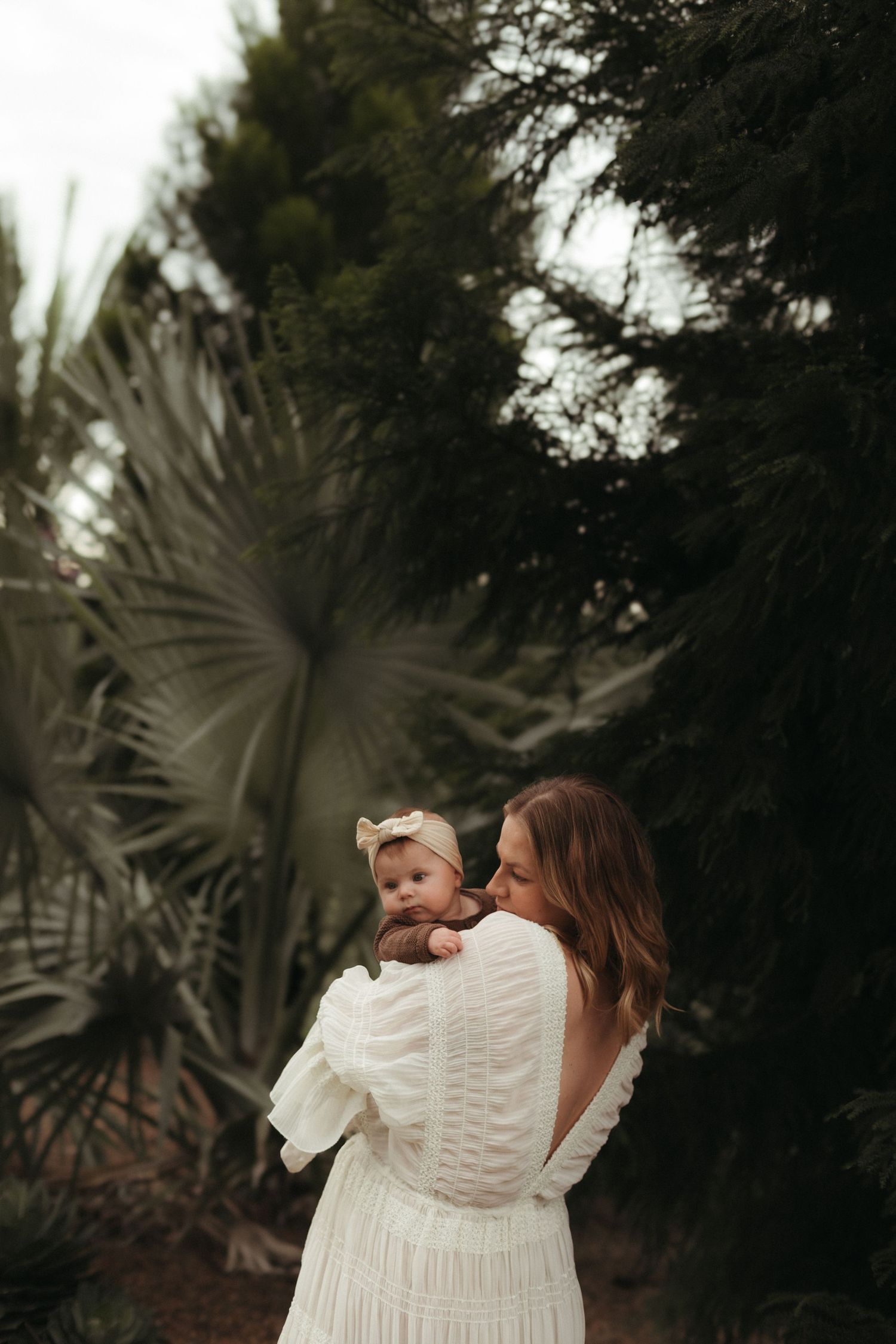 Woman in white dress tenderly kissing baby among tropical palm trees.