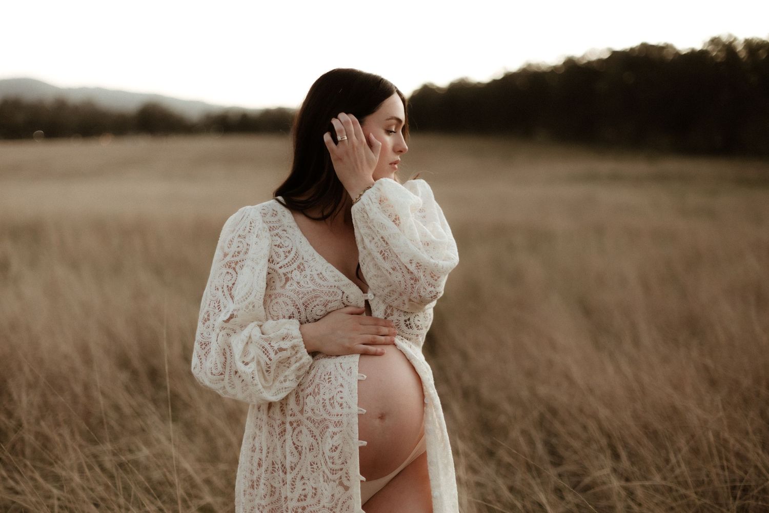 Pregnant woman in white lace robe cradling her baby bump in a golden field at sunset.