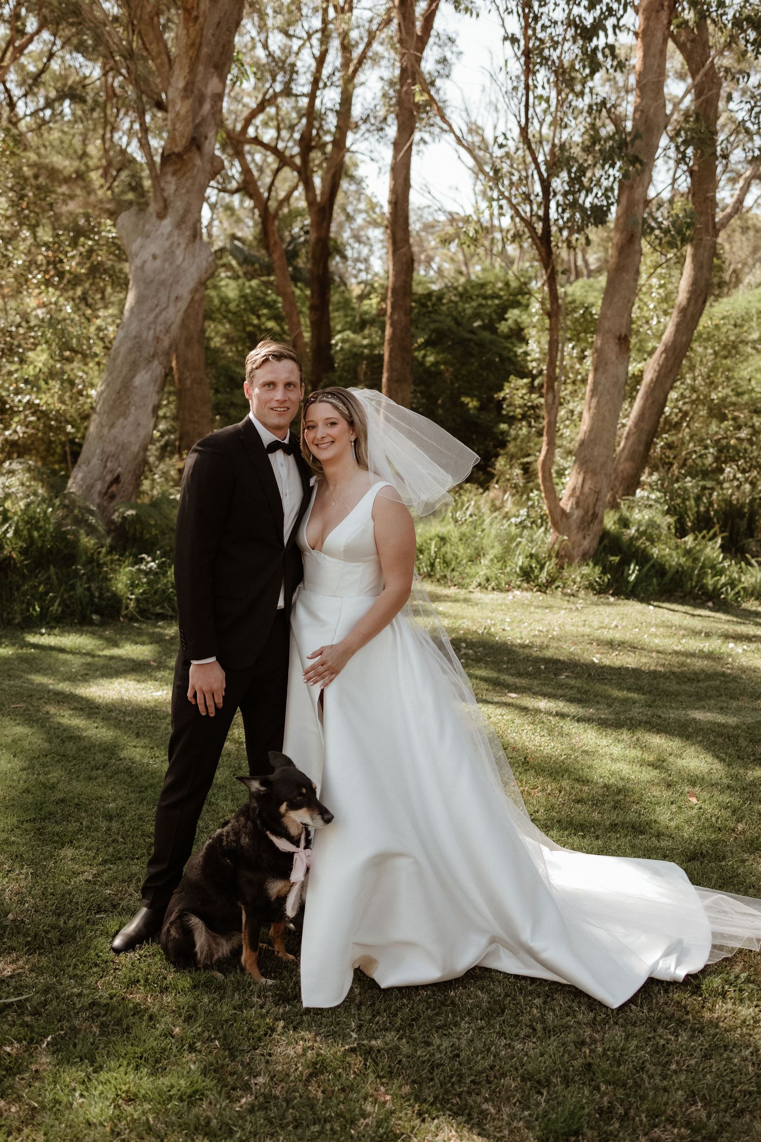 Bride and groom pose with their border collie dog in a beautiful tree-lined garden setting.