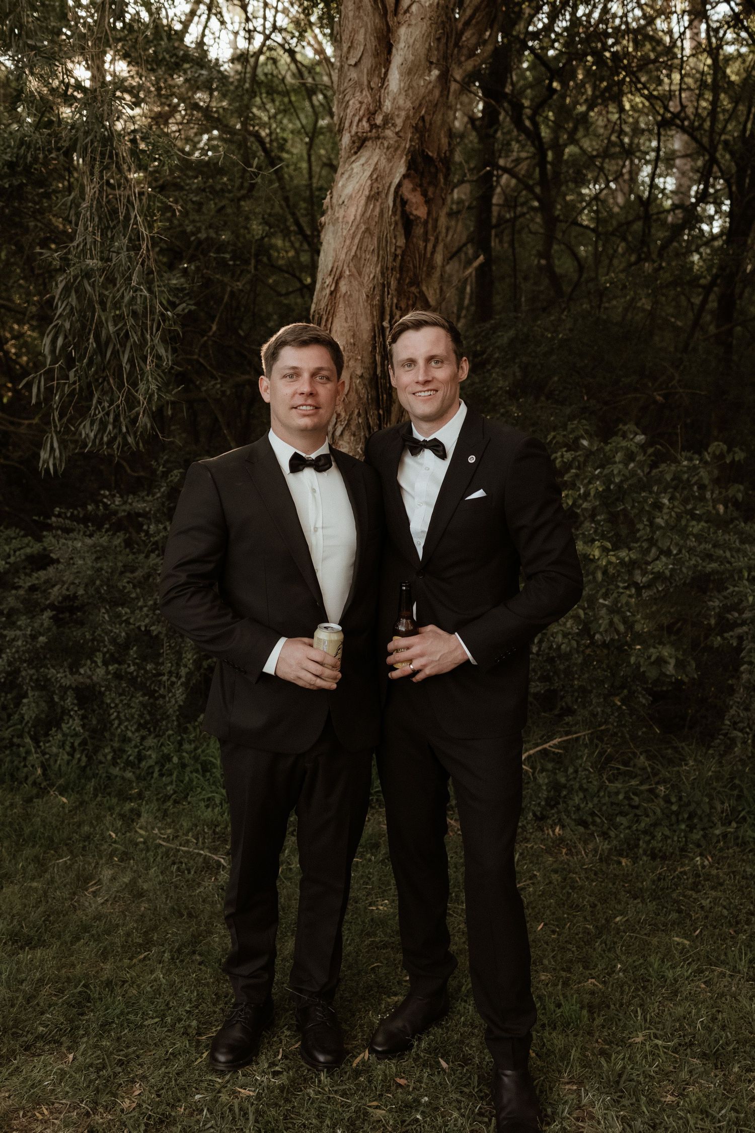 Two men in black tuxedos with bow ties posing together in front of a large tree.