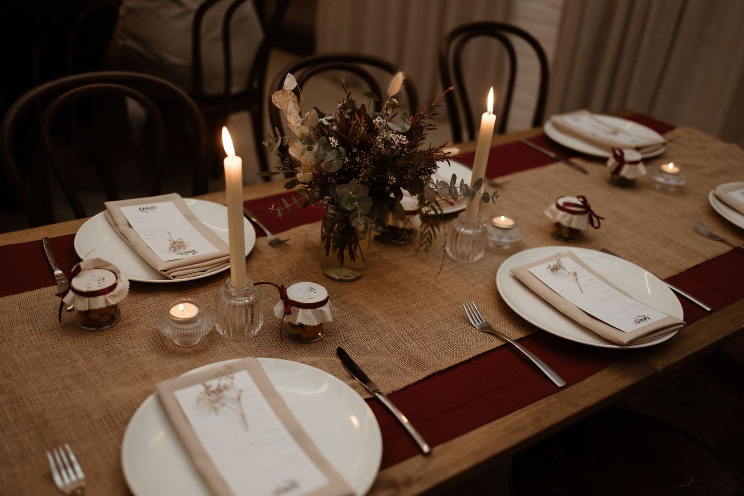 Elegant wedding table setting with burgundy runner, white plates, candles, and floral centerpiece.