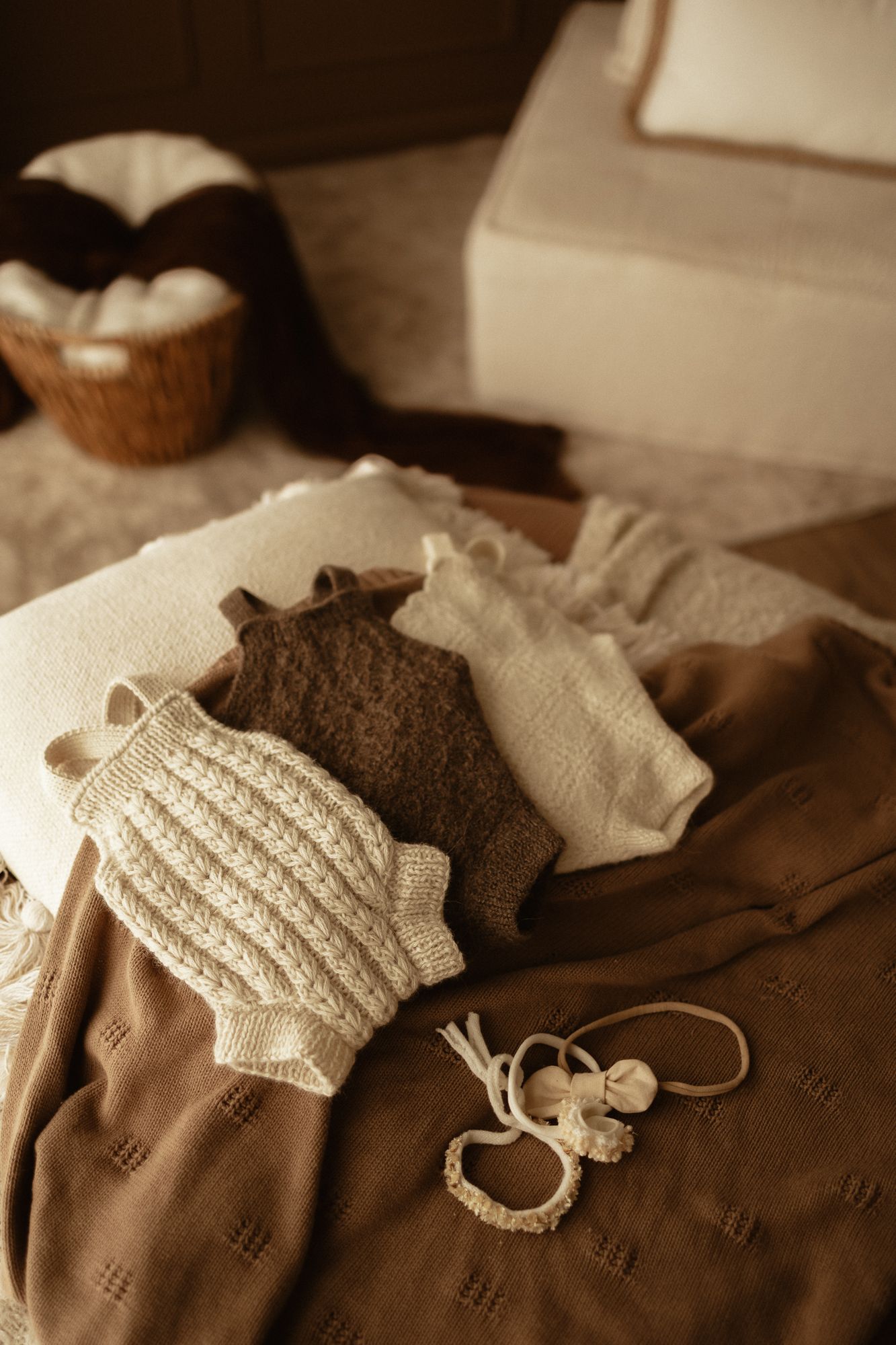 Soft neutral baby clothes including knit onesie and brown blanket arranged on bed with wicker basket.