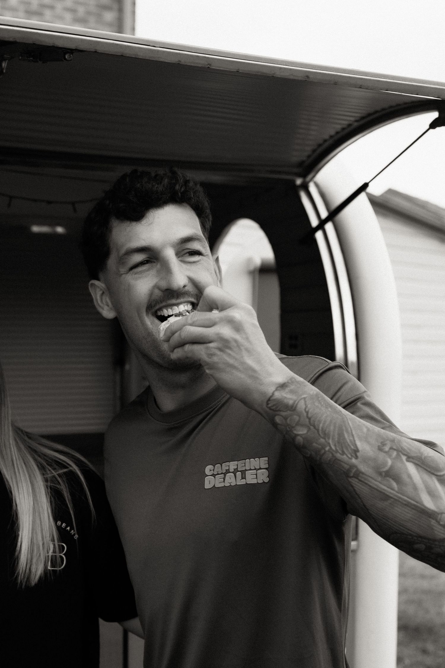 Smiling tattooed man in black Caffeine Dealer shirt standing near a vehicle in black and white photo.
