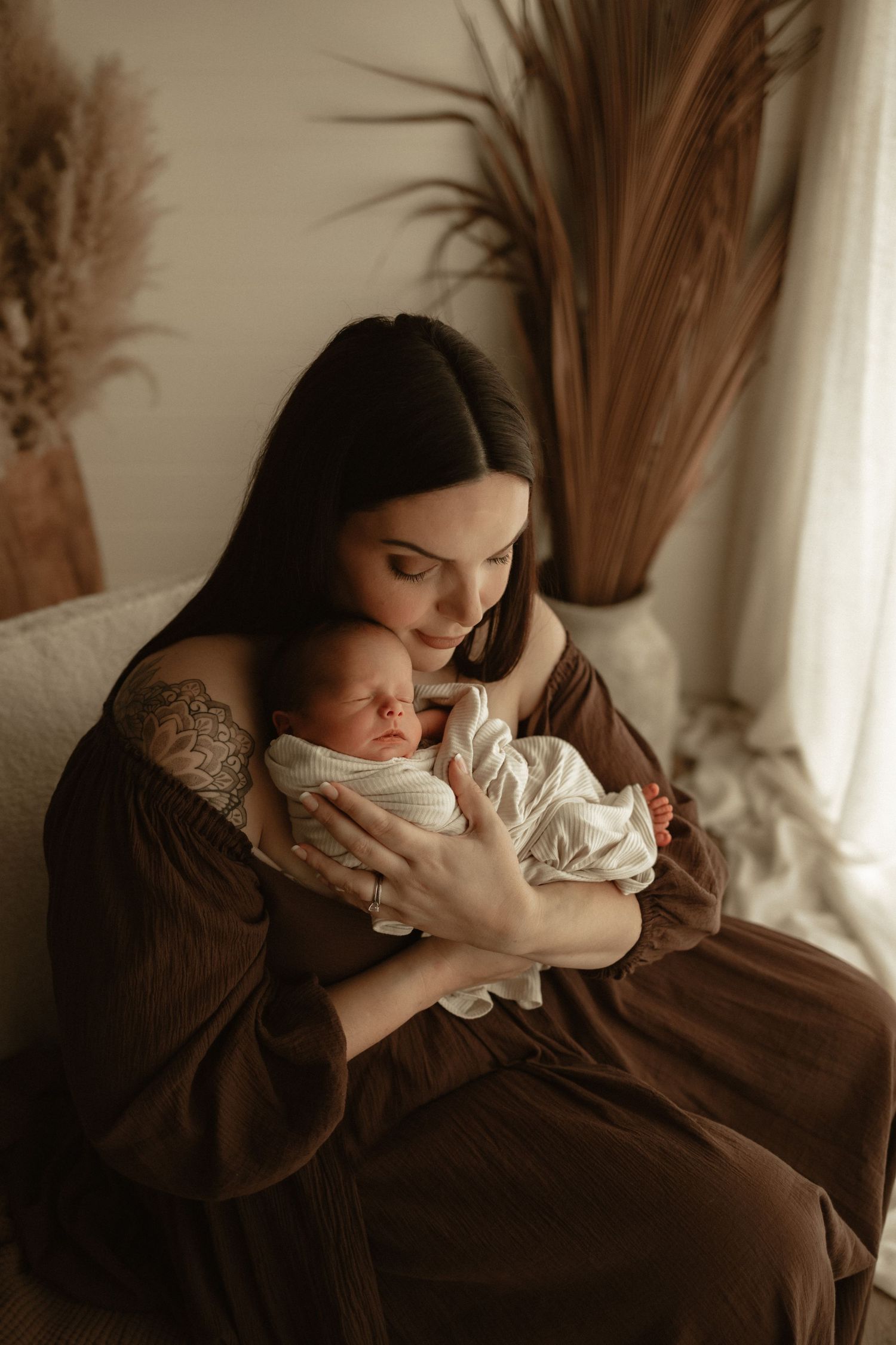 A dark-haired mother in a brown dress tenderly holds her sleeping newborn baby while seated.