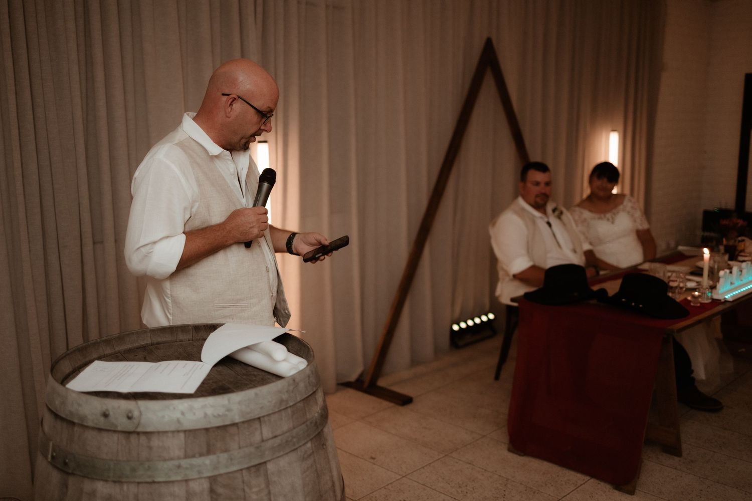 Bald man in white linen suit reads from book while standing at rustic wooden barrel during wedding ceremony.