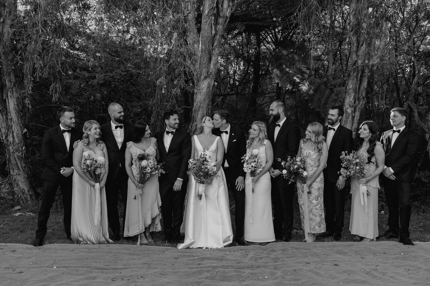 Black and white photo of wedding party laughing together among trees, bridesmaids and groomsmen in formal attire.