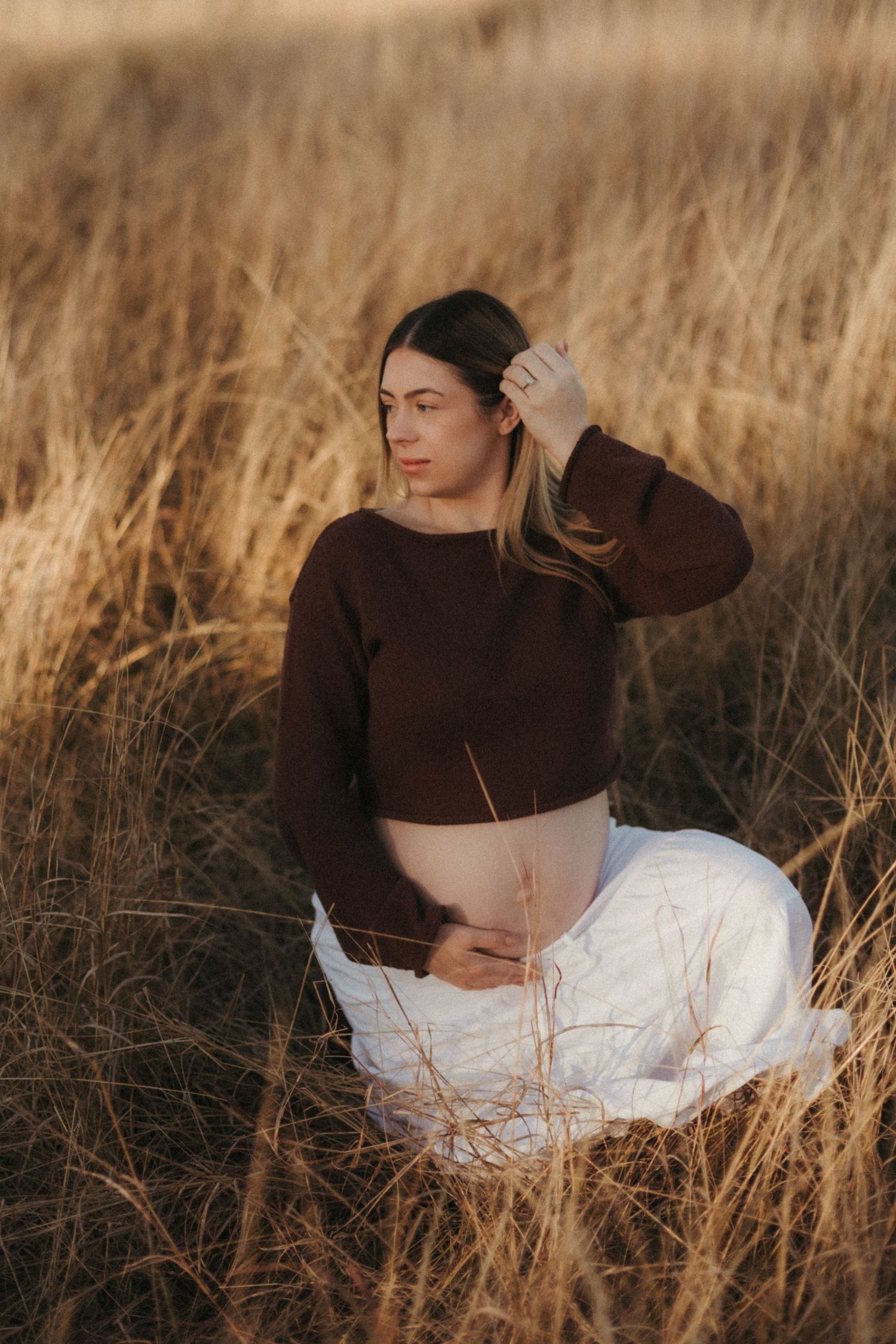 Pregnant woman in brown crop top and white skirt sitting in tall dry grass at golden hour.