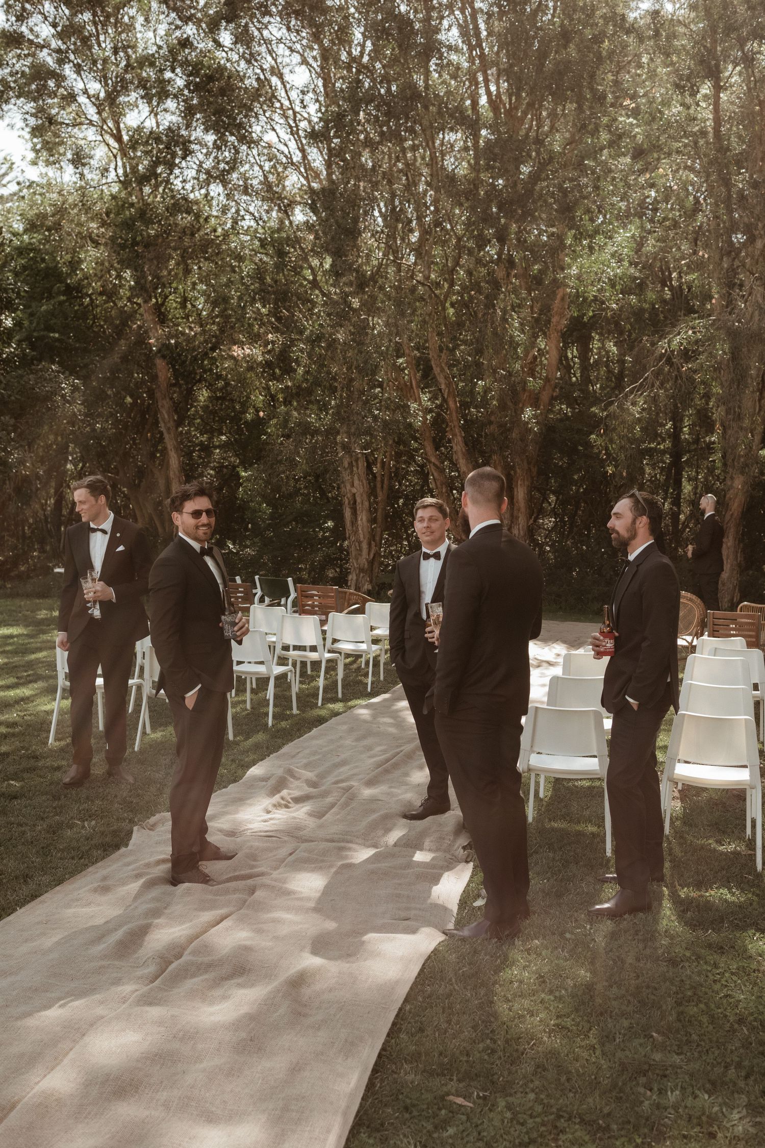 Groomsmen in black tuxedos socializing along the aisle at an outdoor wedding ceremony venue.