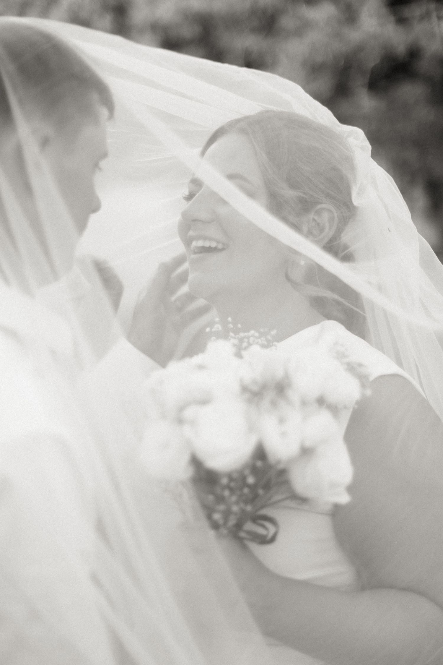 Black and white photo of bride laughing joyfully under flowing veil with bouquet.