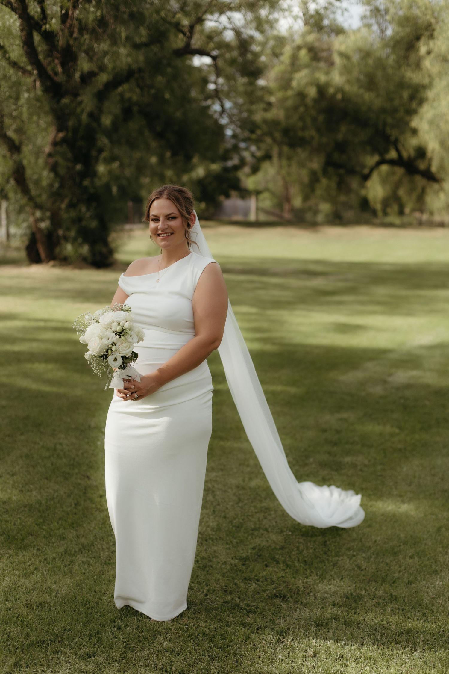 Bride in white one-shoulder gown holding bouquet, standing on green lawn with trees in background.